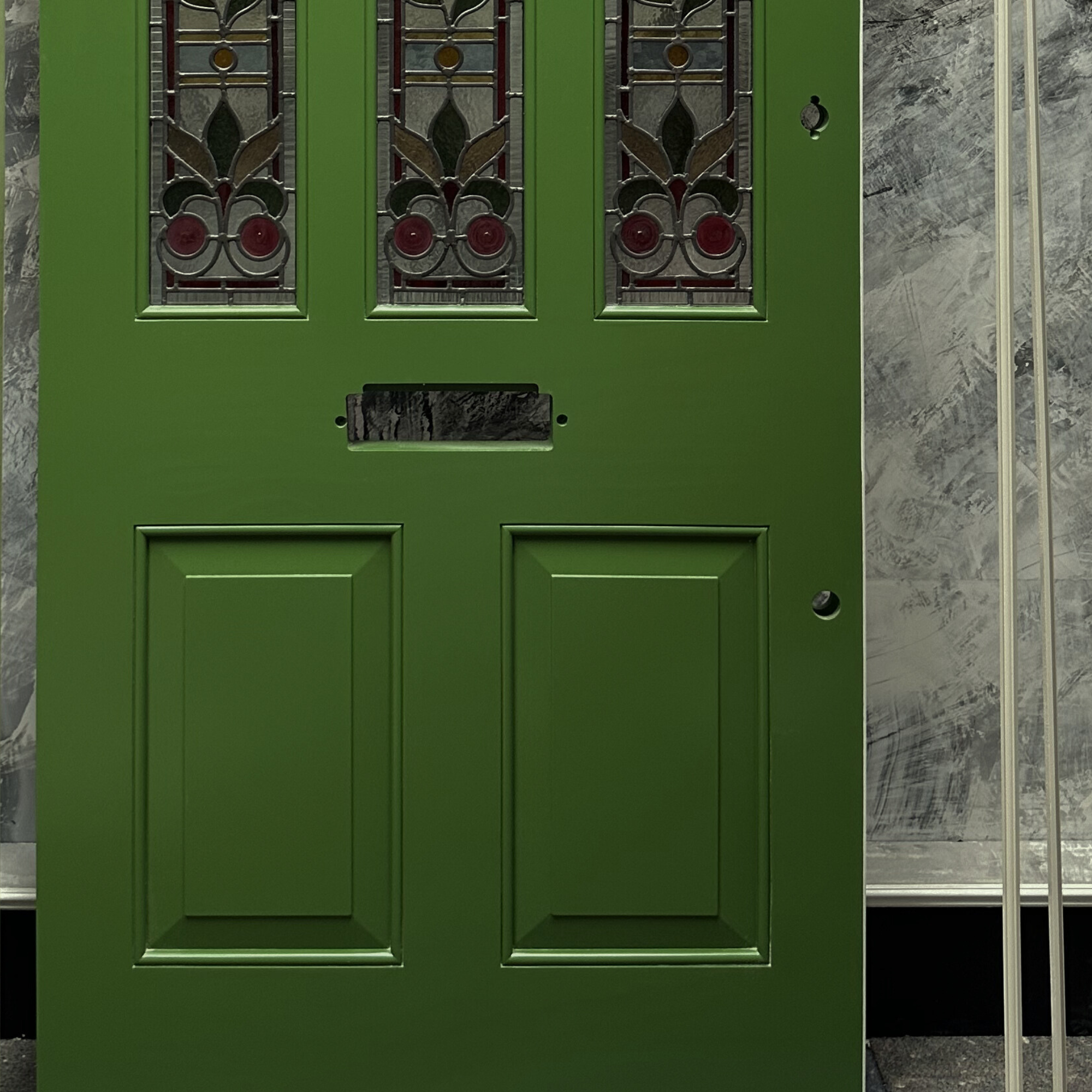 Green door with decorative glass panels at the top, a mail slot in the middle, and a small hole to the right, against a gray textured background. handmade bespoke accoya door.
