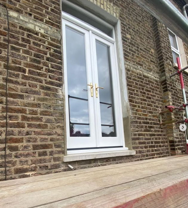 White handmade bespoke accoya door. double-glass door with gold handles on a brick building, with scaffolding on the right side.