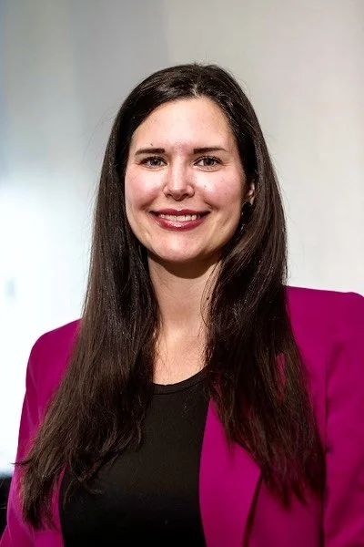 A woman with long dark hair, smiling, wearing a pink blazer and black top, standing against a light-colored background.