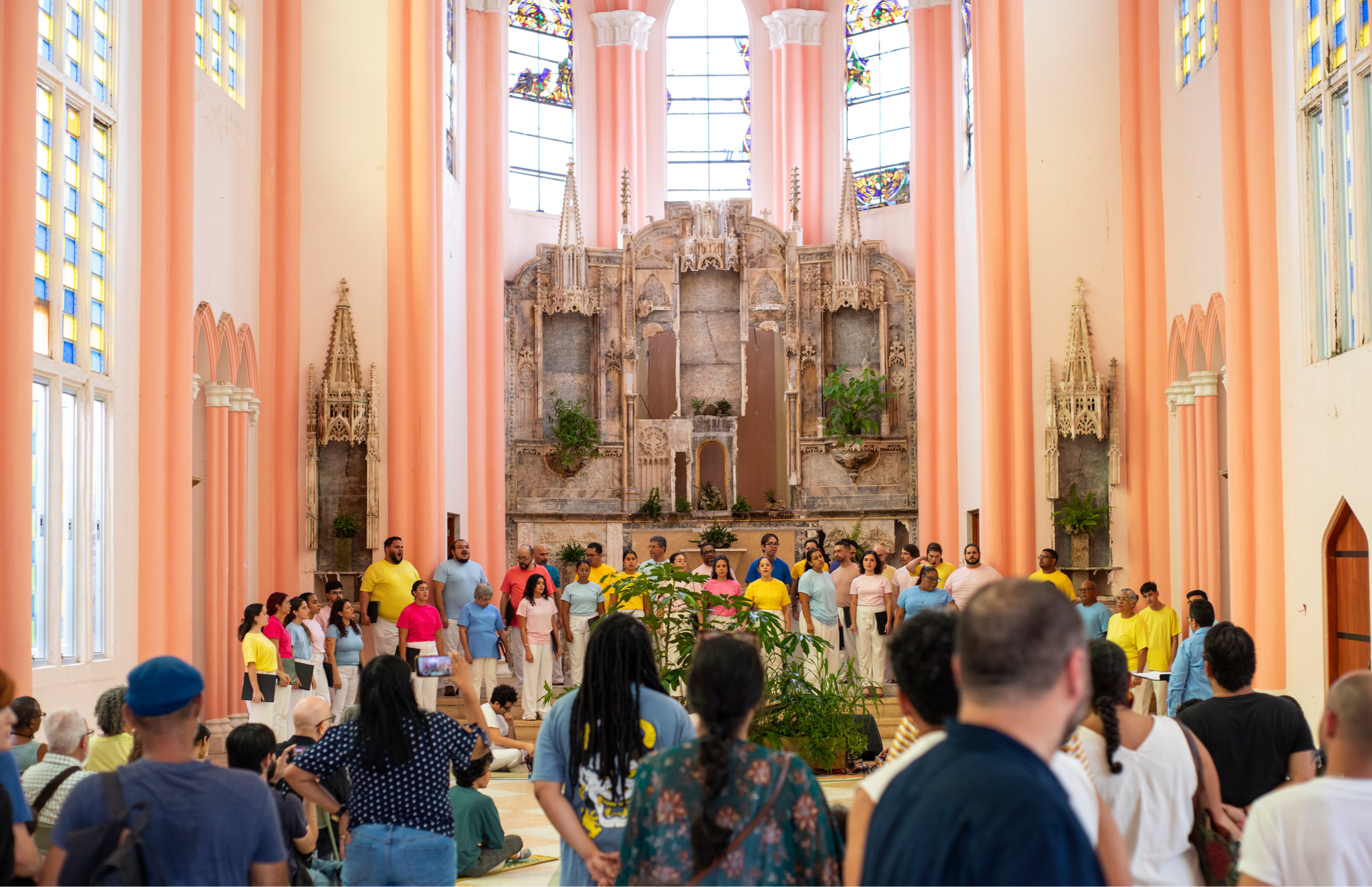 Coro numeroso dispuesto en semicírculo dentro de una iglesia con columnas rosadas y vitrales. Público de pie observa desde el primer plano.