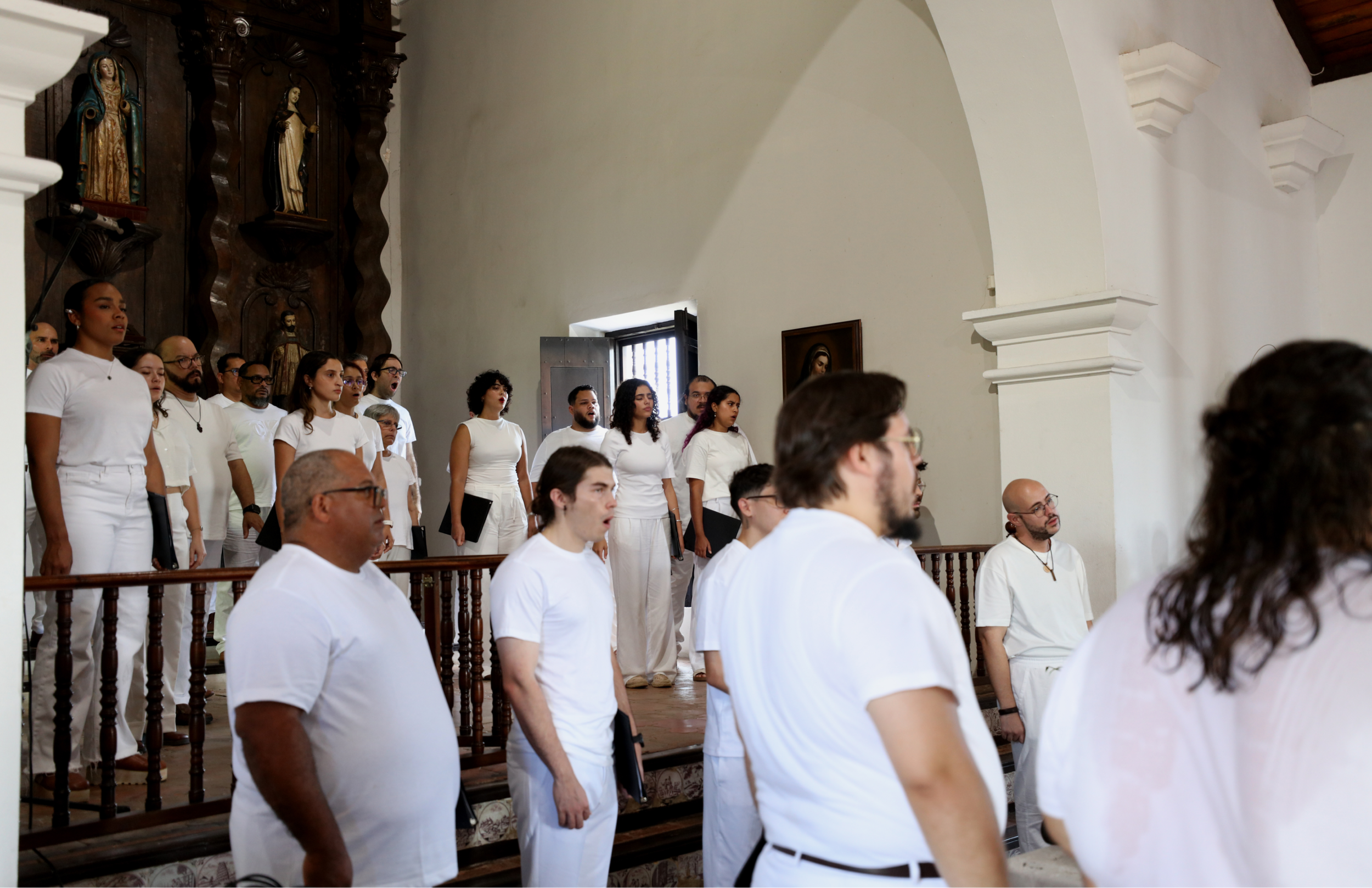 Grupo de cantantes vestidos de blanco de pie junto a un altar tallado en madera. Varias personas en primer plano escuchan mientras sostienen programas o teléfonos.