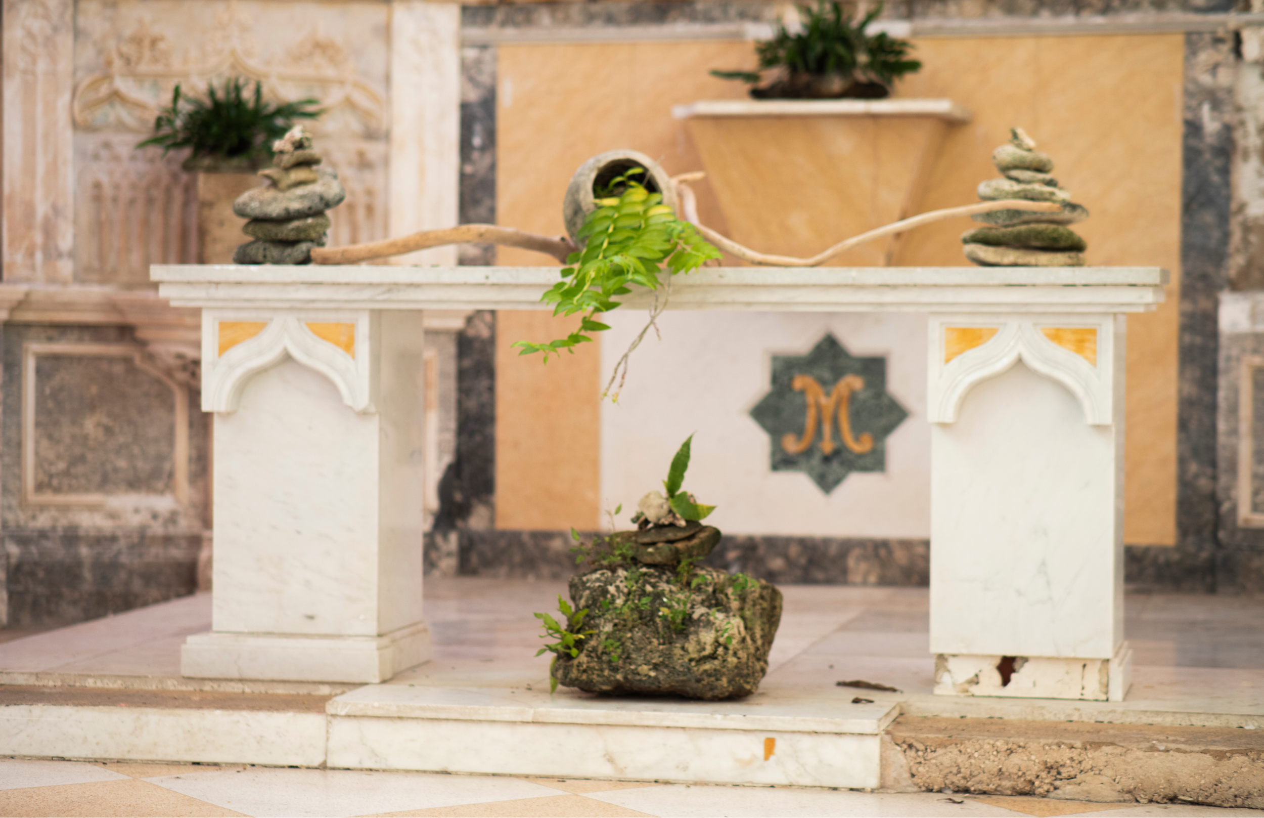 Altar blanco con columnas en el interior de una iglesia. Sobre la mesa hay piedras apiladas, una rama de madera y plantas; en el centro inferior hay una roca con vegetación.