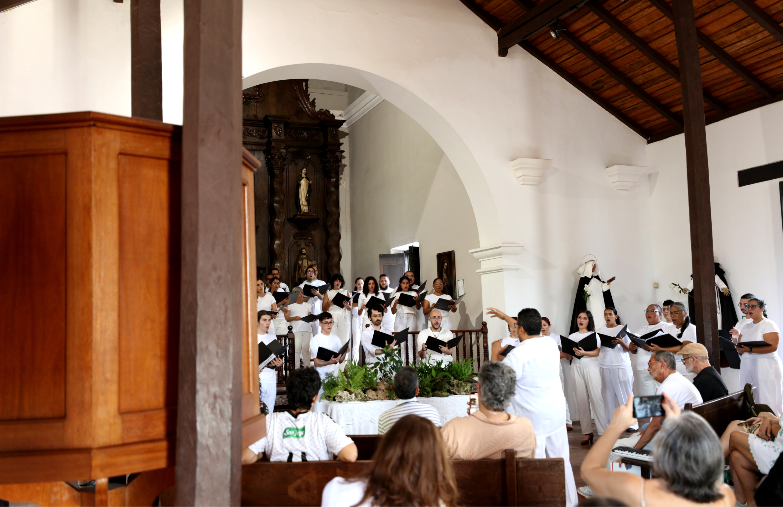 Coro vestido de blanco canta en el interior de la iglesia mientras un director dirige desde el frente. El público observa y algunas personas sostienen teléfonos para grabar.