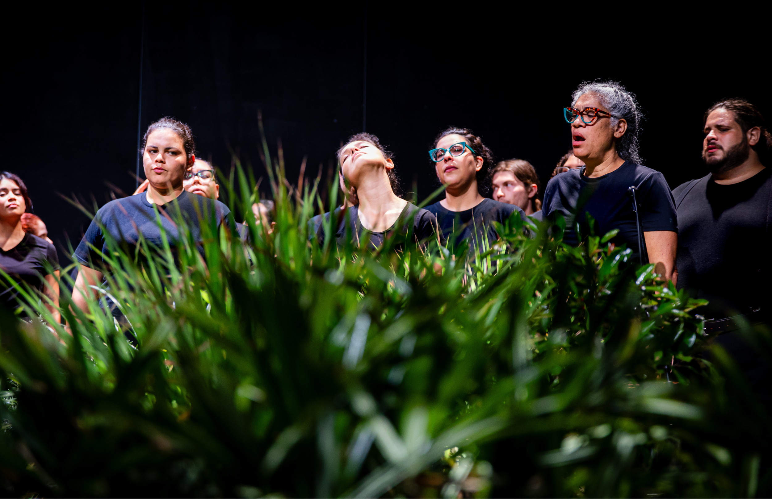 Grupo de cantantes vestidos de negro canta con expresiones intensas, parcialmente cubiertos por plantas en primer plano.