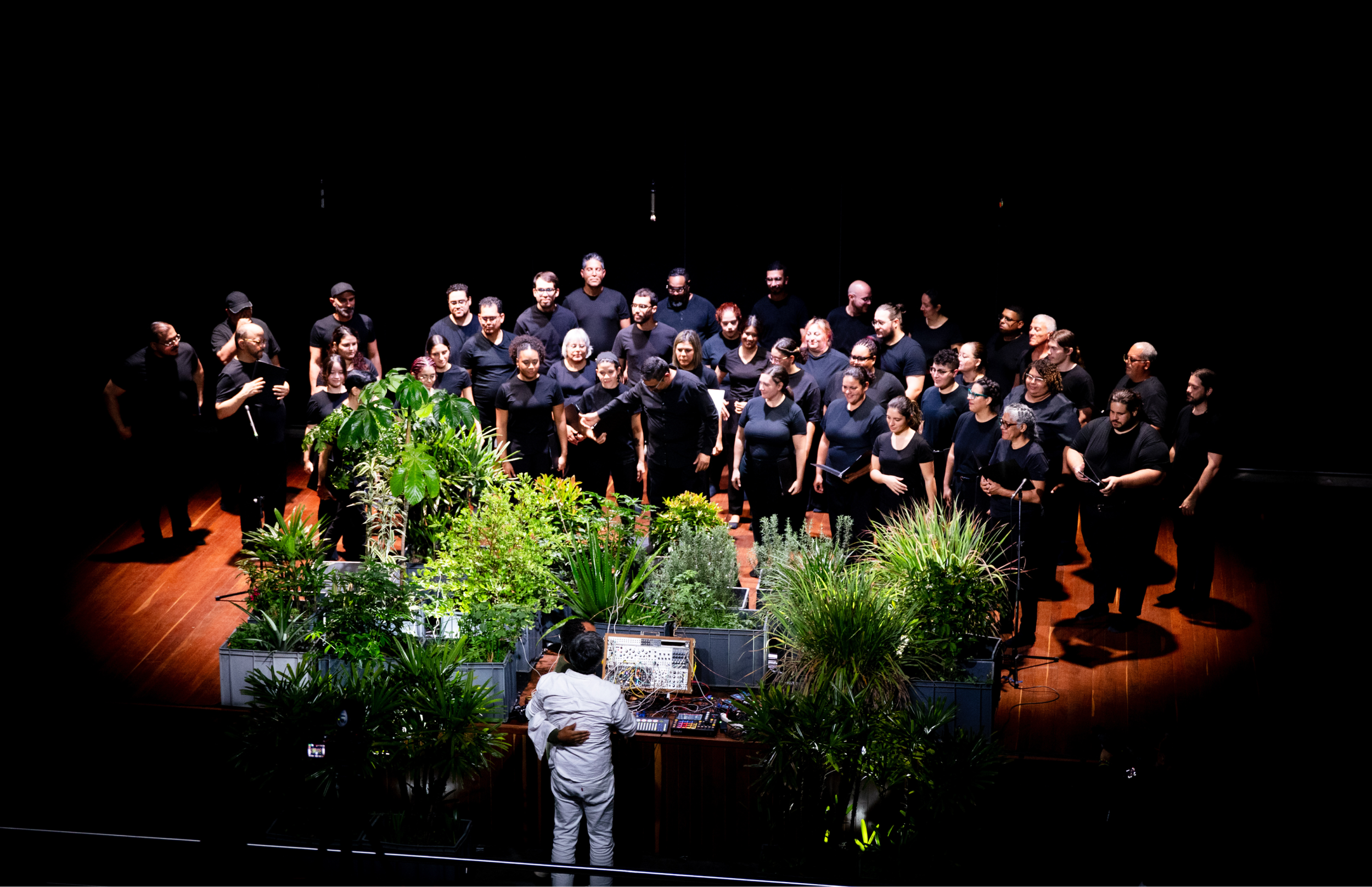 El coro permanece de pie en el escenario después de cantar, rodeando una instalación de plantas. En primer plano, una persona frente a una mesa con equipos electrónicos observa hacia el grupo.