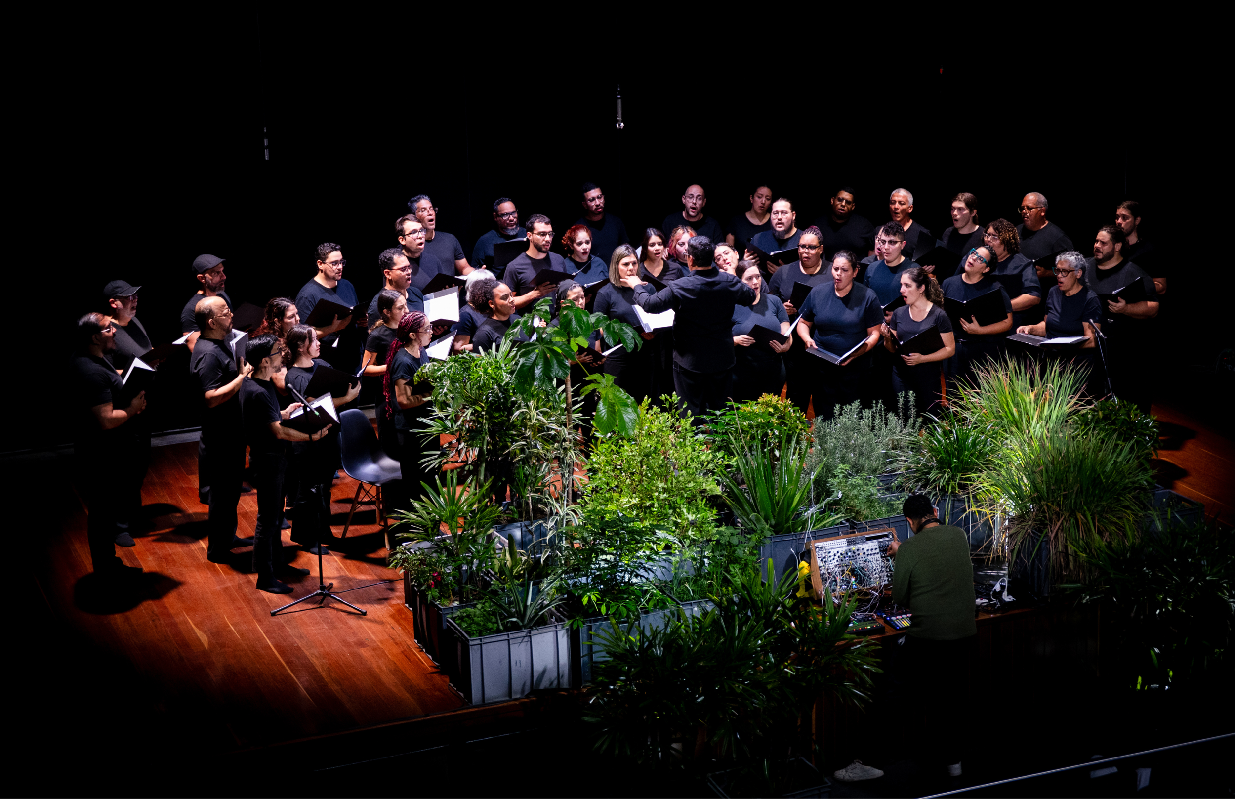 Coro vestido de negro canta en un escenario oscuro alrededor de una instalación central con plantas. Un director dirige al frente mientras una persona opera equipos electrónicos junto a la vegetación.