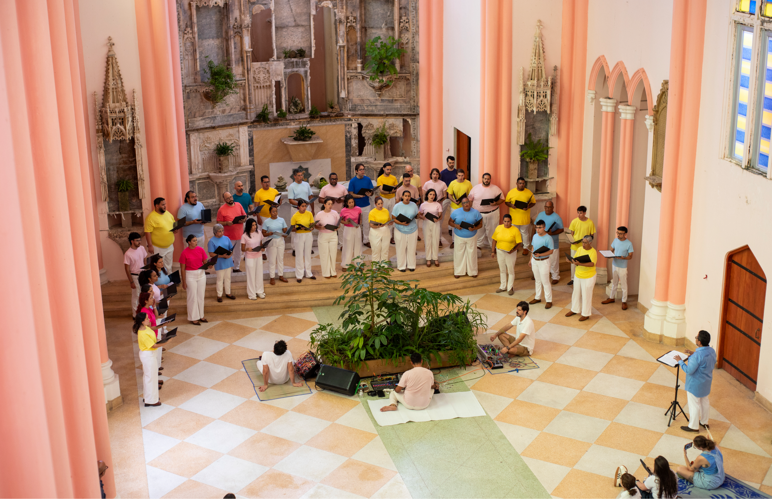 Vista amplia desde arriba de un coro formando un semicírculo alrededor de una instalación vegetal central. Varias personas están sentadas en el suelo junto a equipos electrónicos mientras el público observa alrededor.