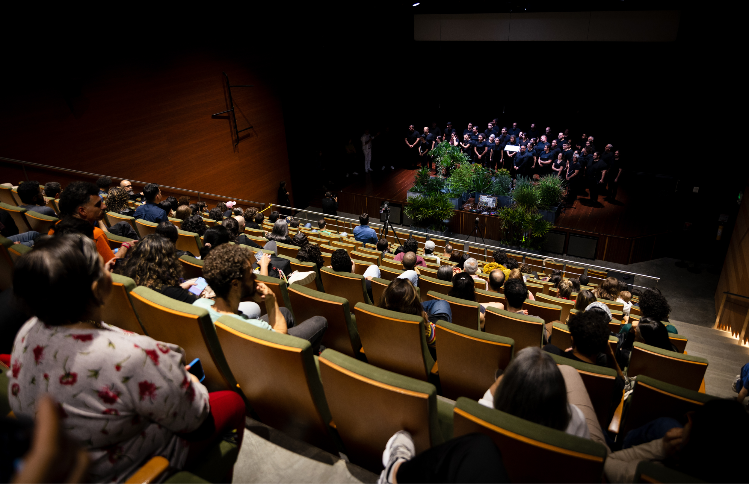 Vista general del escenario con iluminación puntual sobre el coro vestido de negro. La instalación vegetal ocupa el centro mientras una persona opera equipos electrónicos al frente.