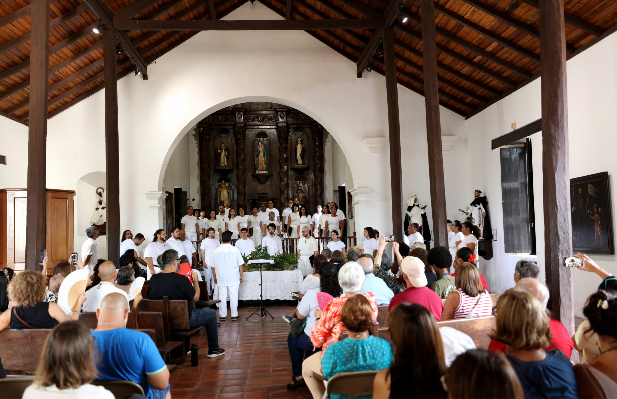 Vista amplia del interior de la iglesia con techo de madera. Un coro vestido de blanco canta frente a un altar tallado mientras el público observa desde las bancas.