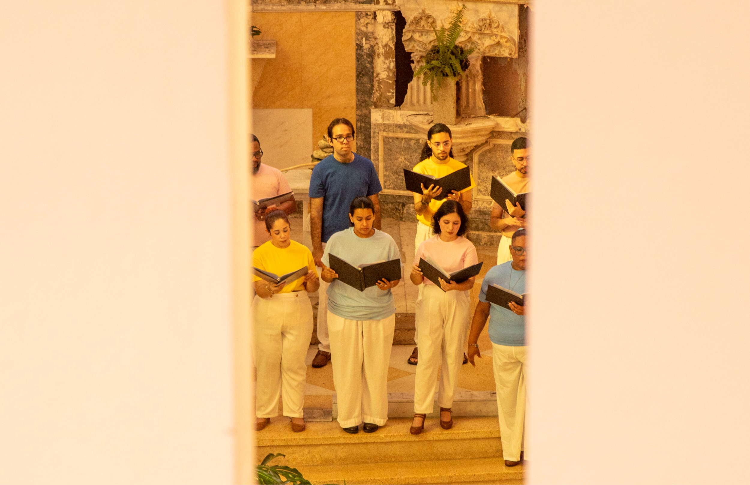 Grupo de personas de pie en las escalinatas de una iglesia, sosteniendo carpetas negras y cantando. Visten camisetas en colores pastel y pantalones blancos; detrás se ve un altar con plantas.
