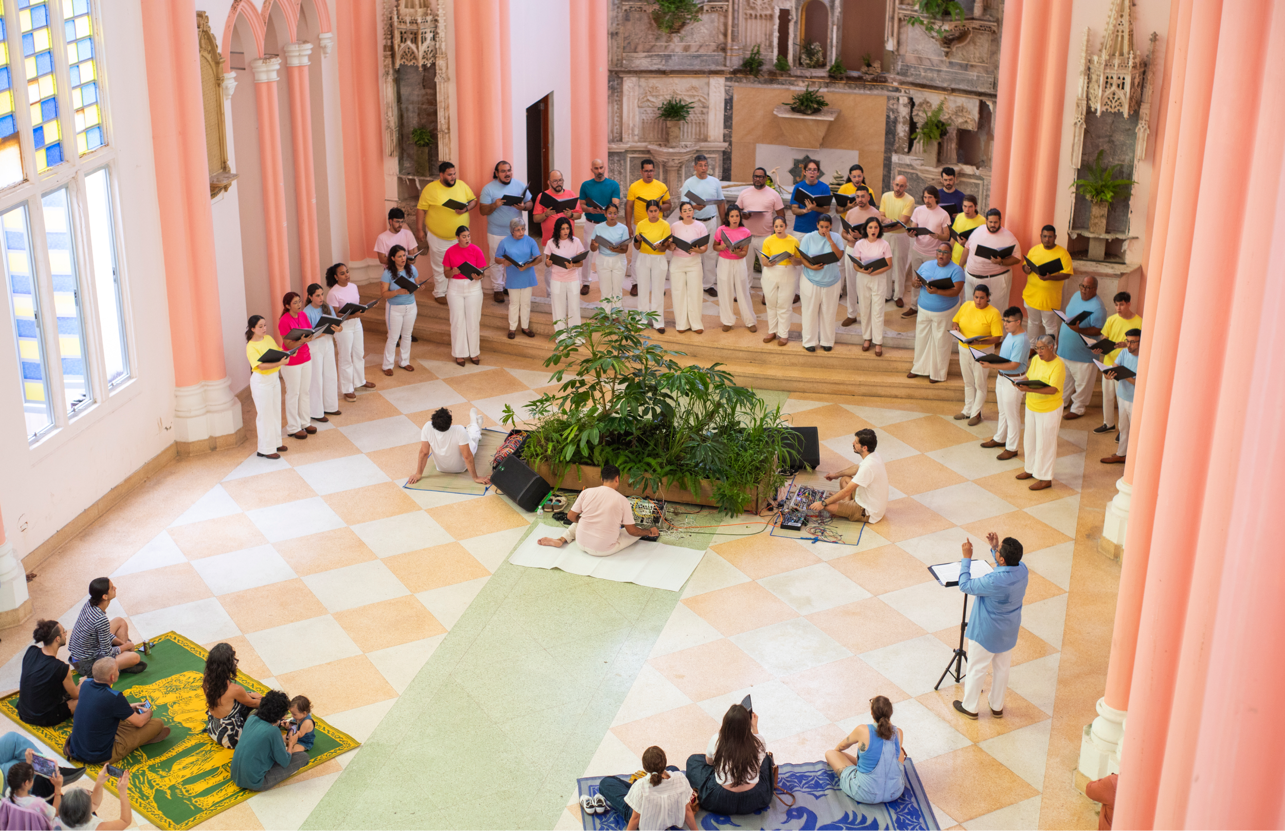 Vista cenital amplia del interior de la iglesia. El coro rodea una instalación de plantas en el centro mientras músicos sentados operan dispositivos electrónicos; el director dirige desde un atril.