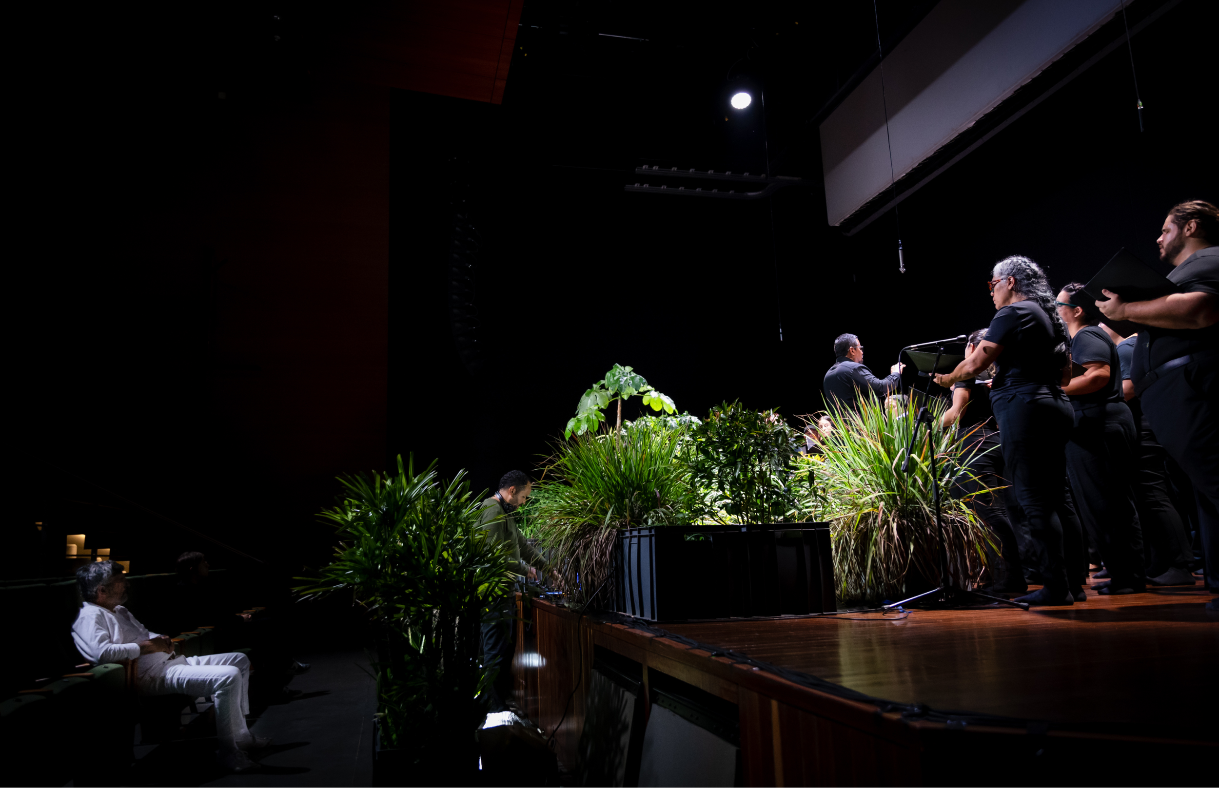 Vista amplia del teatro desde las butacas. El público observa al coro en el escenario, dispuesto alrededor de una instalación central de plantas.