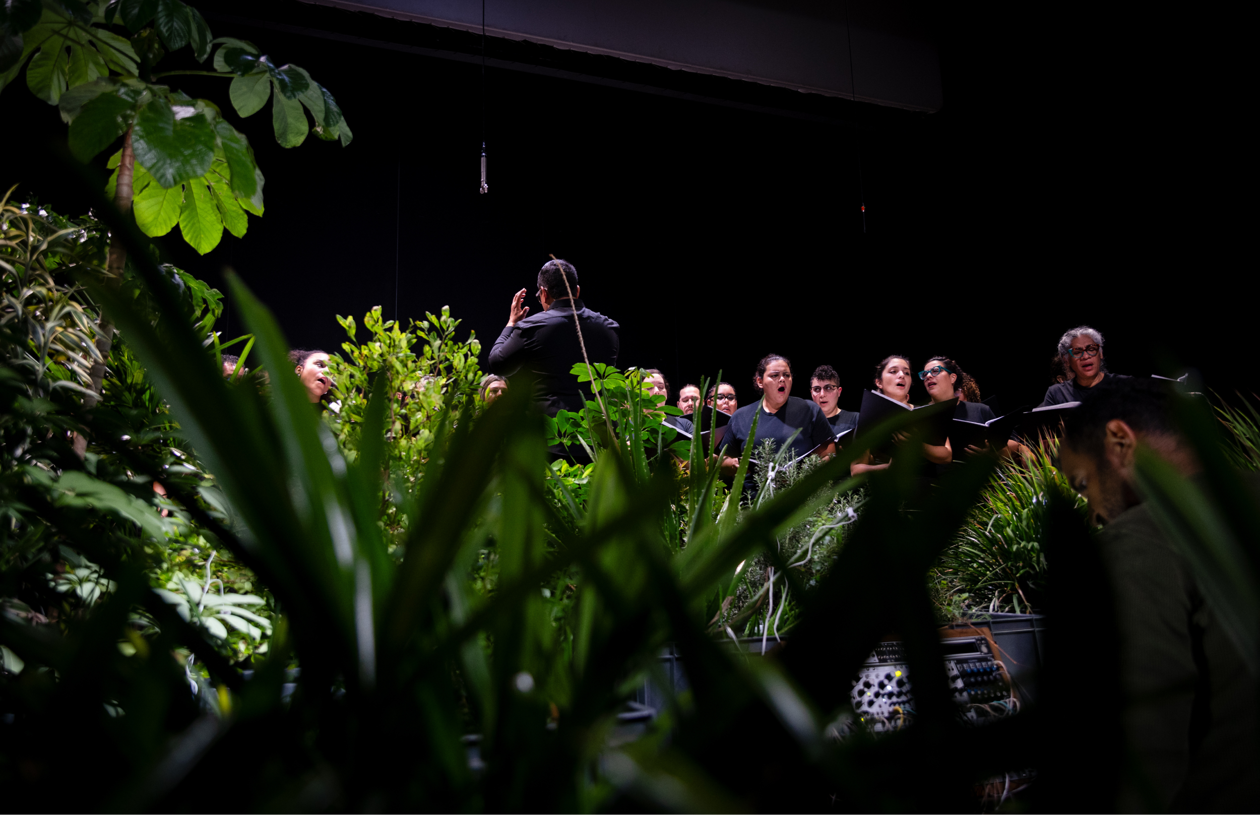 Vista lateral del escenario: el director dirige al coro mientras una persona ajusta equipos electrónicos junto a la instalación de plantas. Parte del público es visible en la oscuridad.