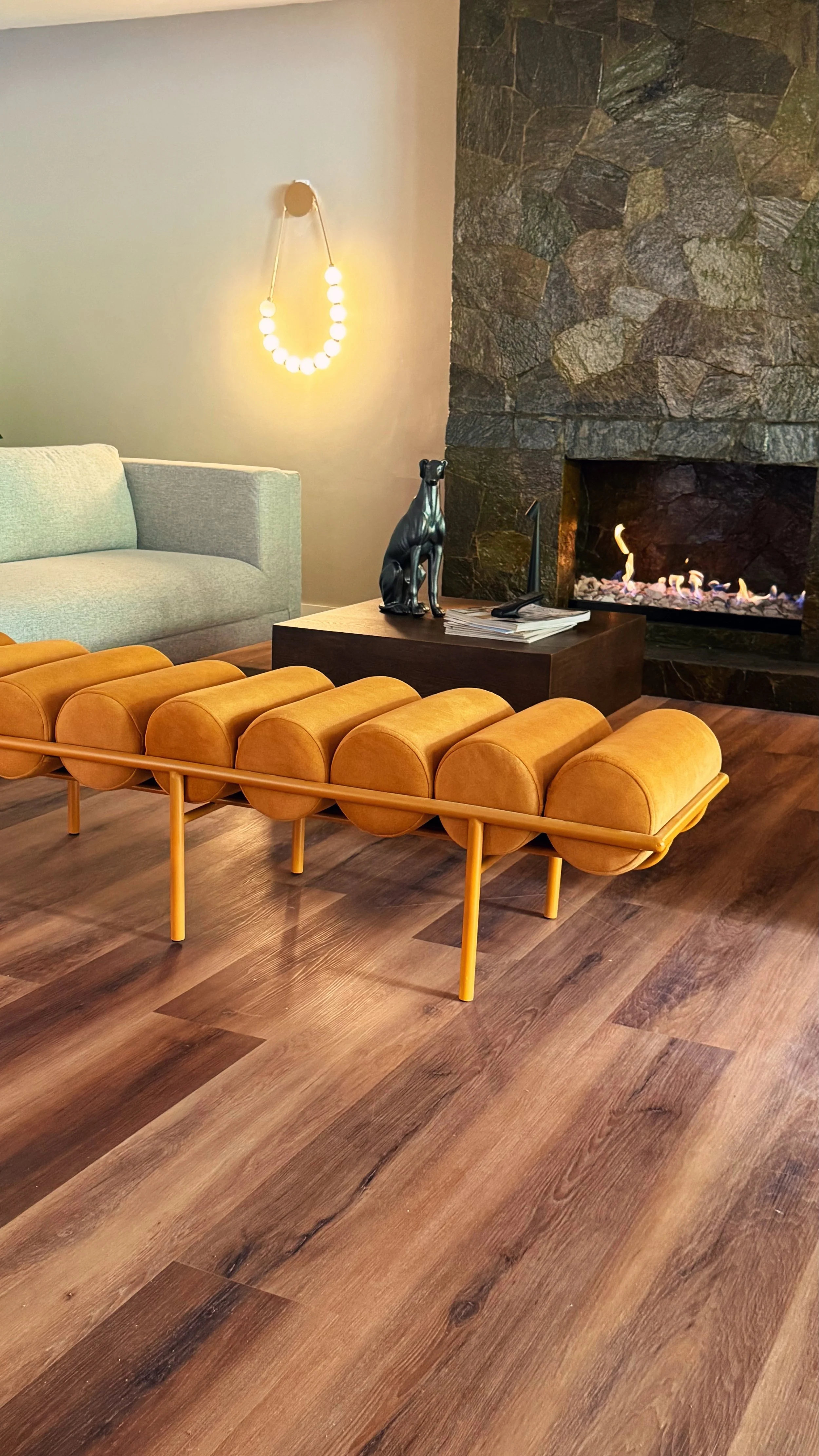 Living room with a green couch, orange bench, black dog sculpture, fireplace, and wall light with a semi-circle of bulbs.