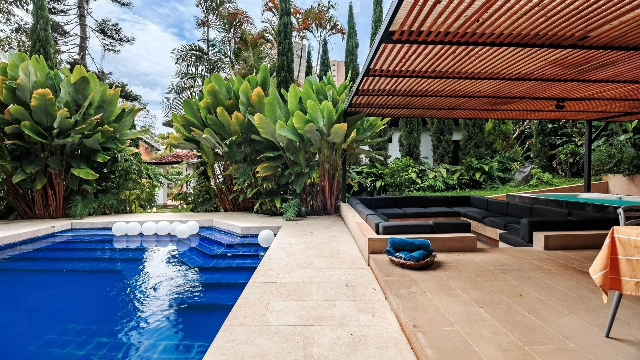An outdoor patio area featuring a swimming pool with blue tiles and floating white balls, surrounded by lush green plants, tall trees, and a covered seating area with black cushioned furniture and a hot tub, under a wooden slat roof.