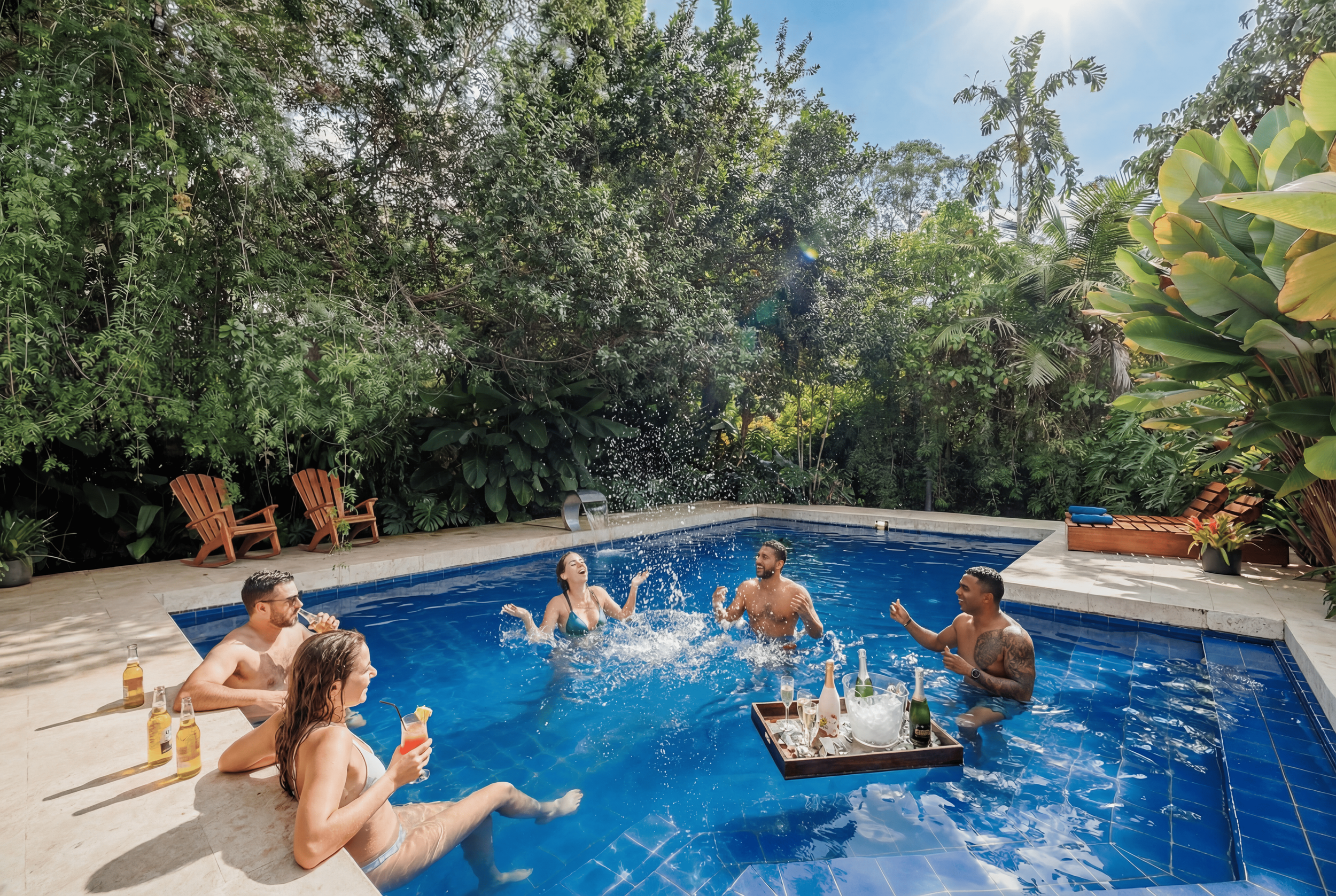 People enjoying a pool party on a sunny day surrounded by lush green trees, with some holding drinks, and others splashing in the pool.