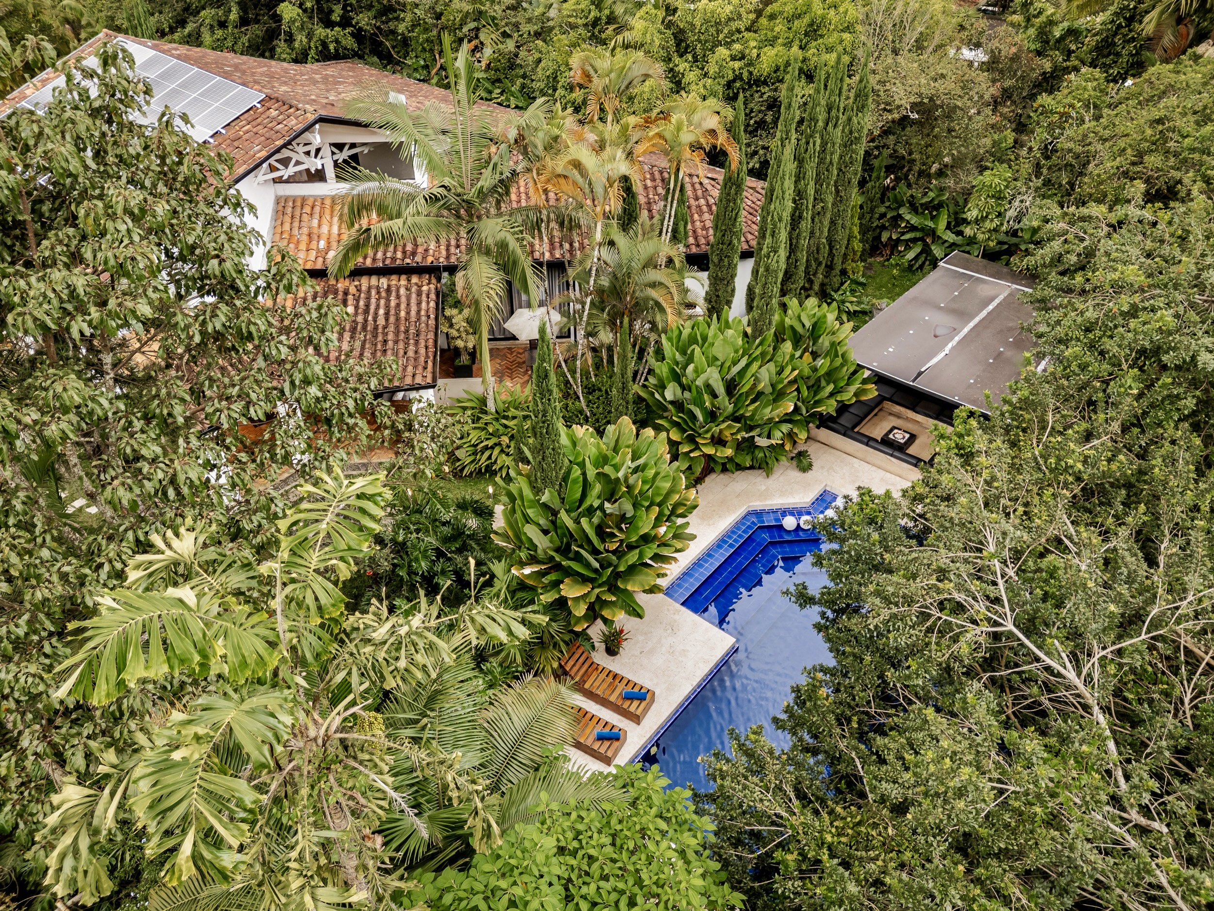 Aerial view of a house with a tiled roof and solar panels, surrounded by lush green trees and plants, with a swimming pool in the yard.