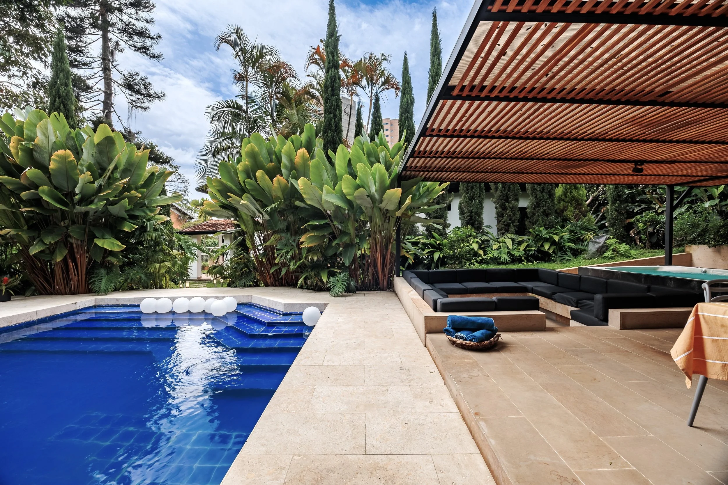 Outdoor swimming pool with blue tiles and floating white spheres, surrounded by beige stone patio, lush green plants and tall trees, with modern furniture and a wooden pergola providing shade.