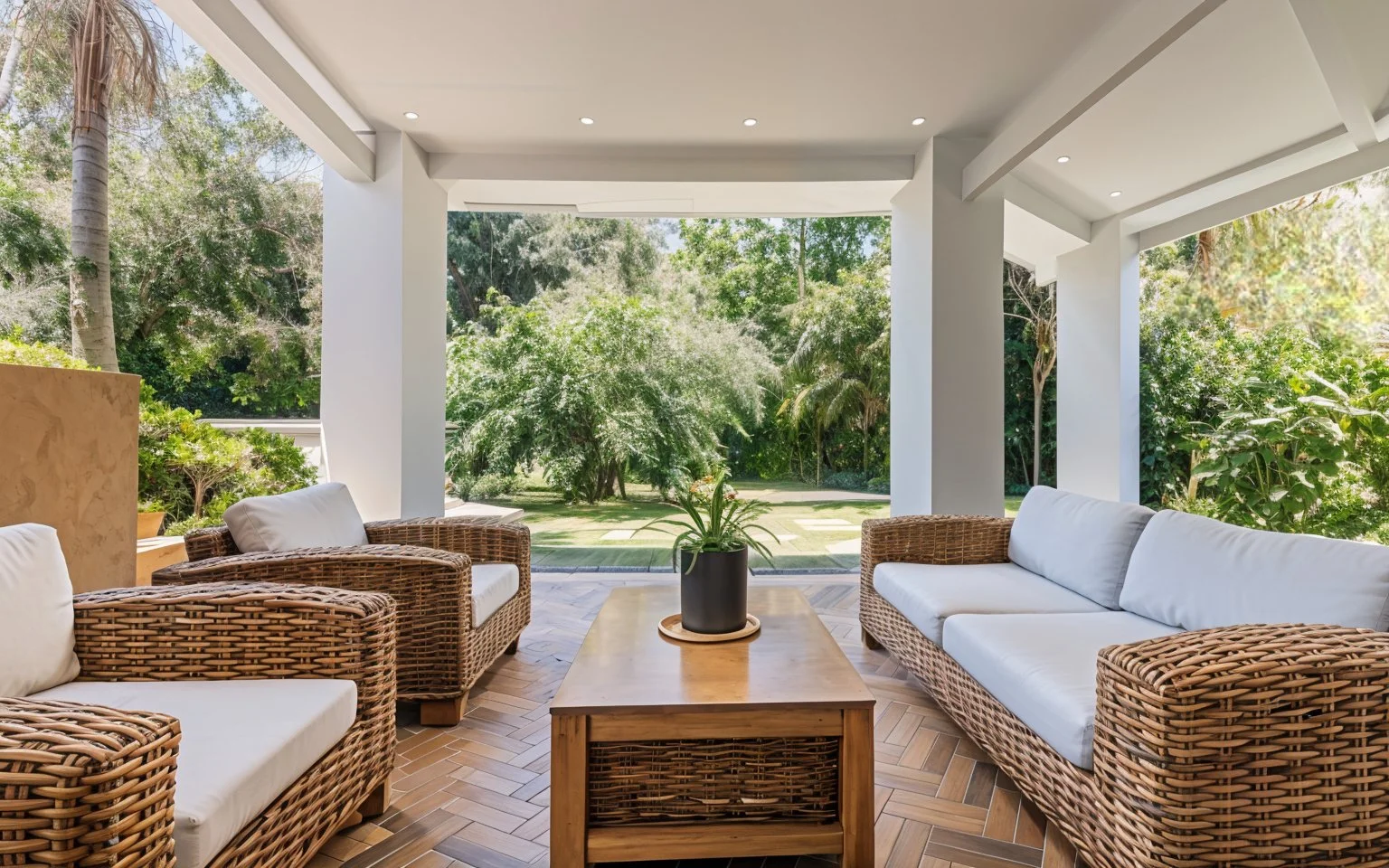 Covered patio with white cushions on wicker furniture, including two armchairs and a sofa, and a wooden coffee table with a potted plant, overlooking a lush green garden.