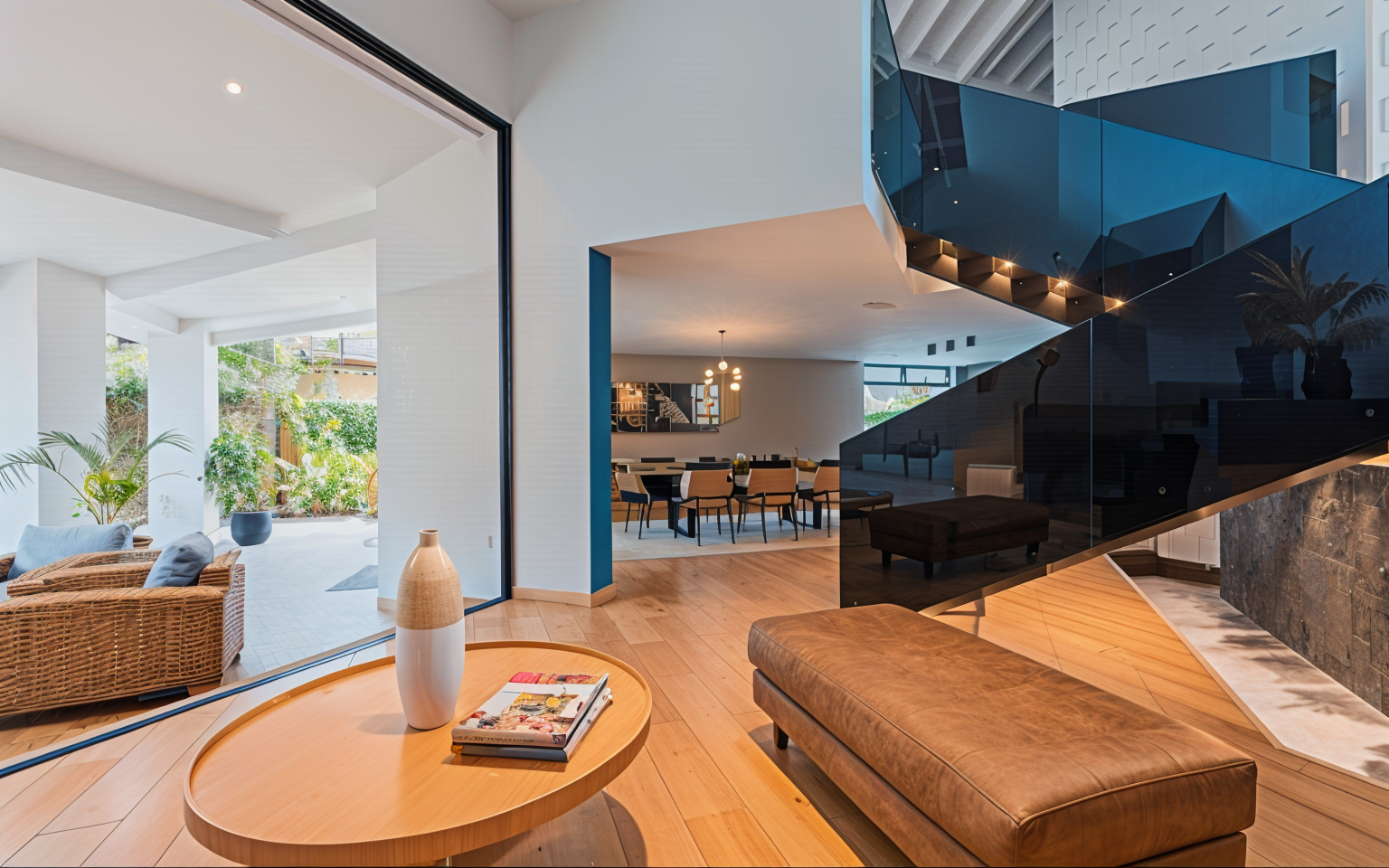 Open-concept living and dining area with wooden flooring, modern furniture, and a large staircase with reflective blue glass panels. The space includes a lounging area with a table and a milk-shaped vase, and large sliding glass doors opening to a pa