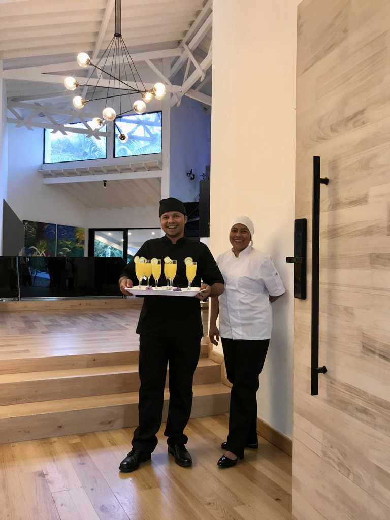 Two restaurant staff members, a man and a woman, standing indoors; the man is holding a tray with four yellow drinks garnished with lemon slices, and they are smiling.