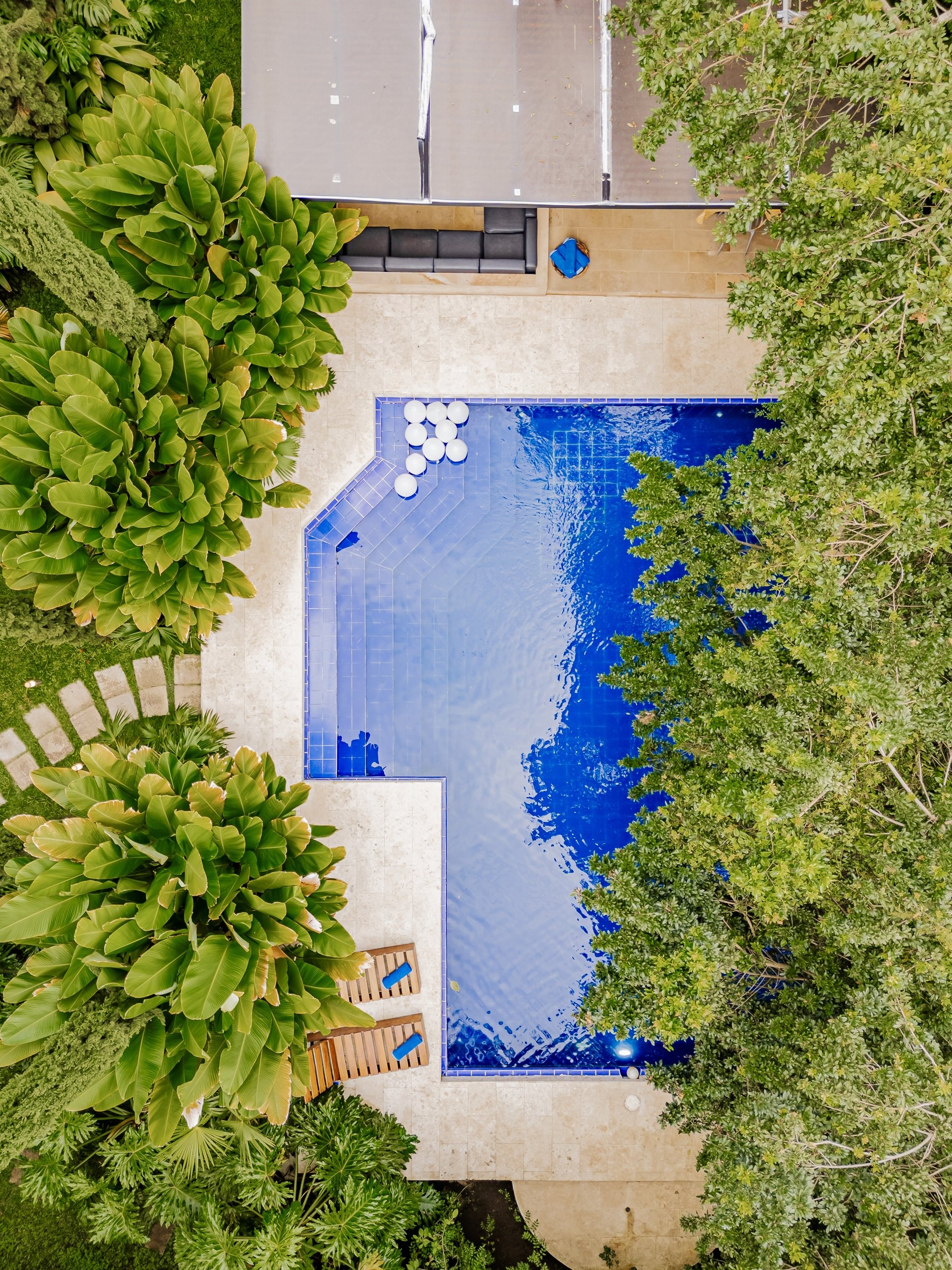A view from above of a swimming pool surrounded by lush green trees and plants, with lounge chairs and a patio area.
