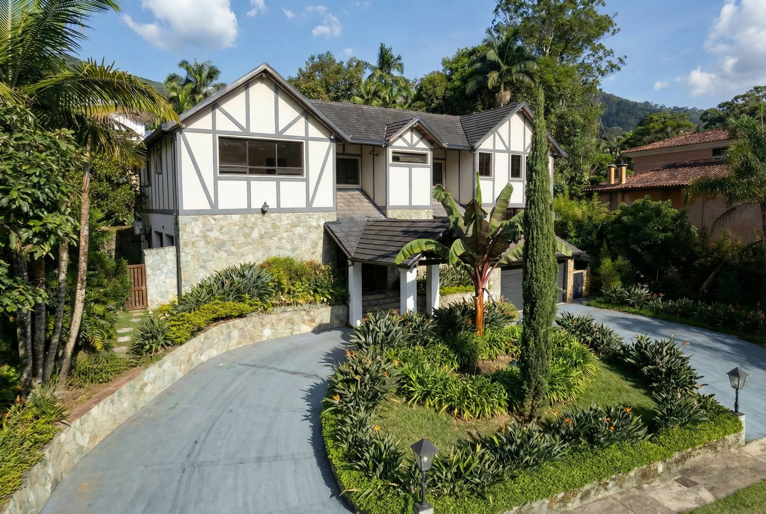 A two-story house with Tudor-style white exterior and black trim, stone foundation, surrounded by lush tropical plants, with a curved driveway and lamp posts, under a blue sky with scattered clouds.