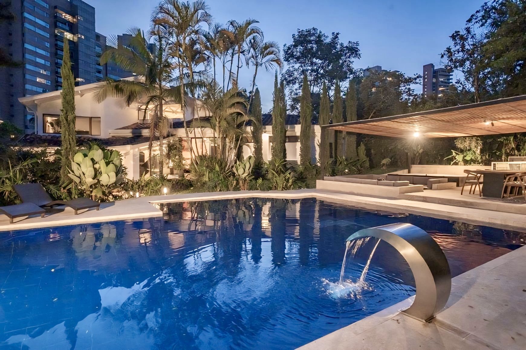 A luxurious modern outdoor pool area during evening with a waterfall feature, surrounded by tropical plants, tall trees, and urban buildings. There are poolside lounge chairs, a shaded seating area with sofas, and a dining table with chairs, all illu