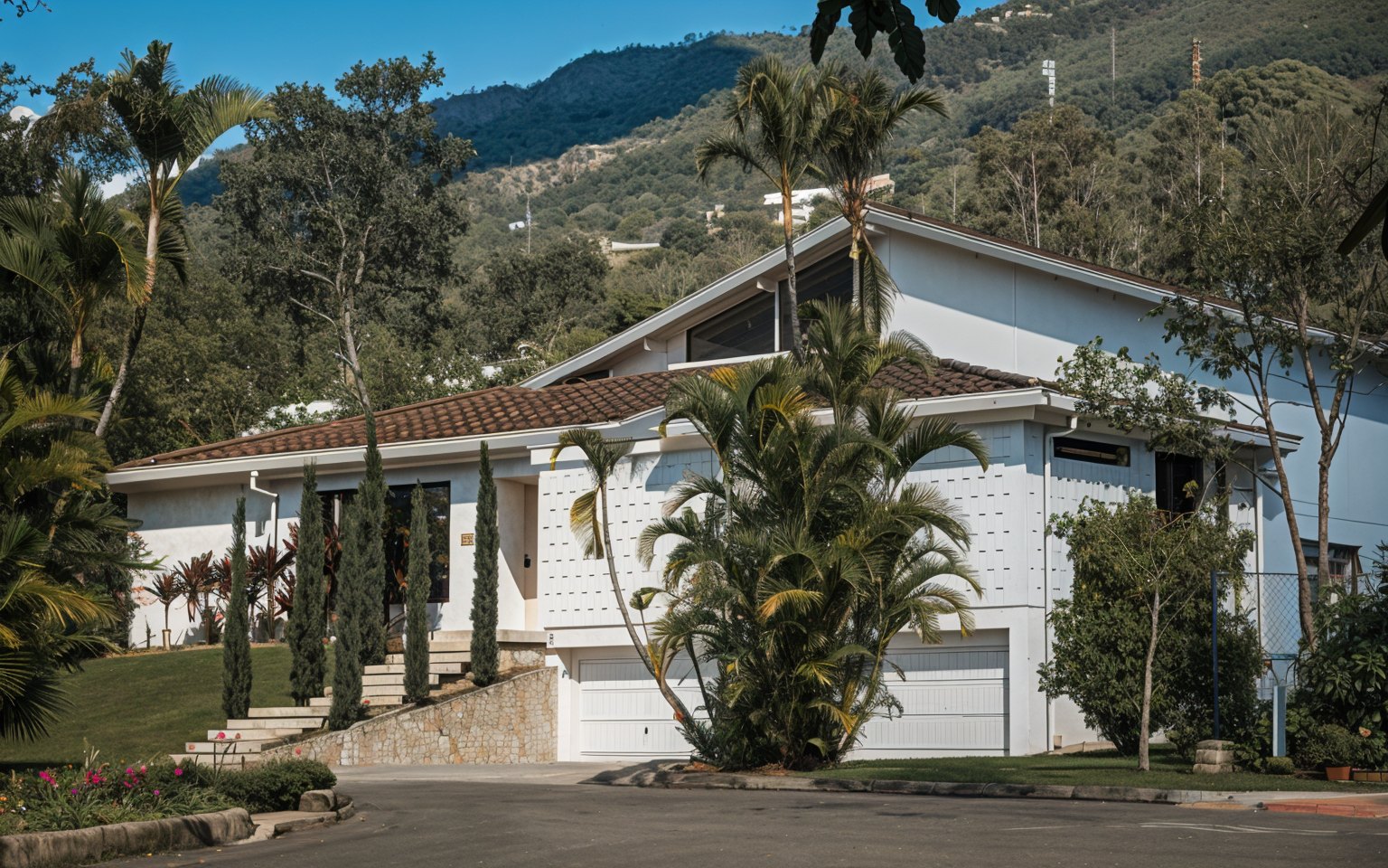 A modern white house with a brown tiled roof, surrounded by palm trees and other greenery, set against a backdrop of green mountains under a clear blue sky.