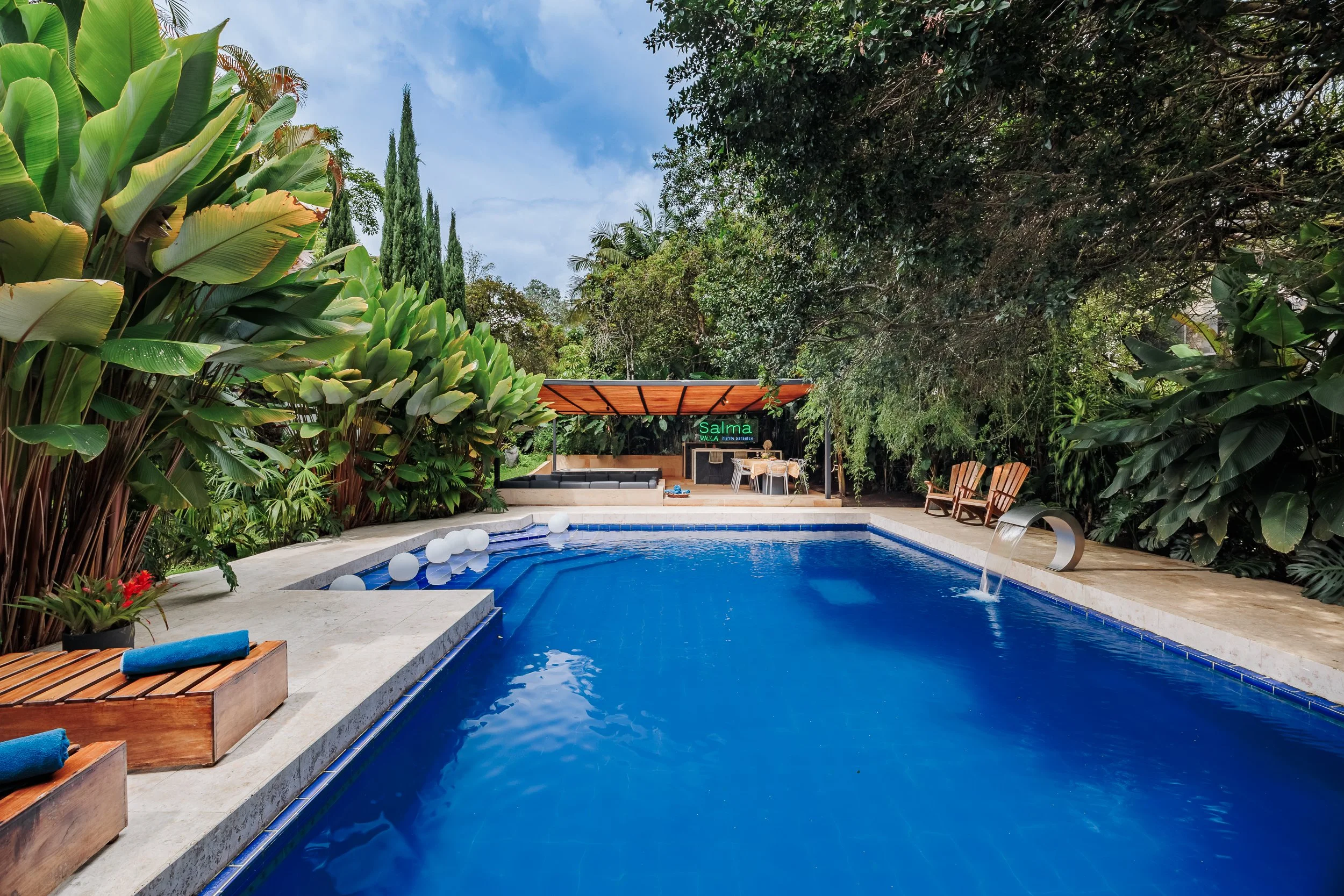A backyard pool surrounded by lush green tropical plants and trees, with a shaded seating area and outdoor dining table in the background.