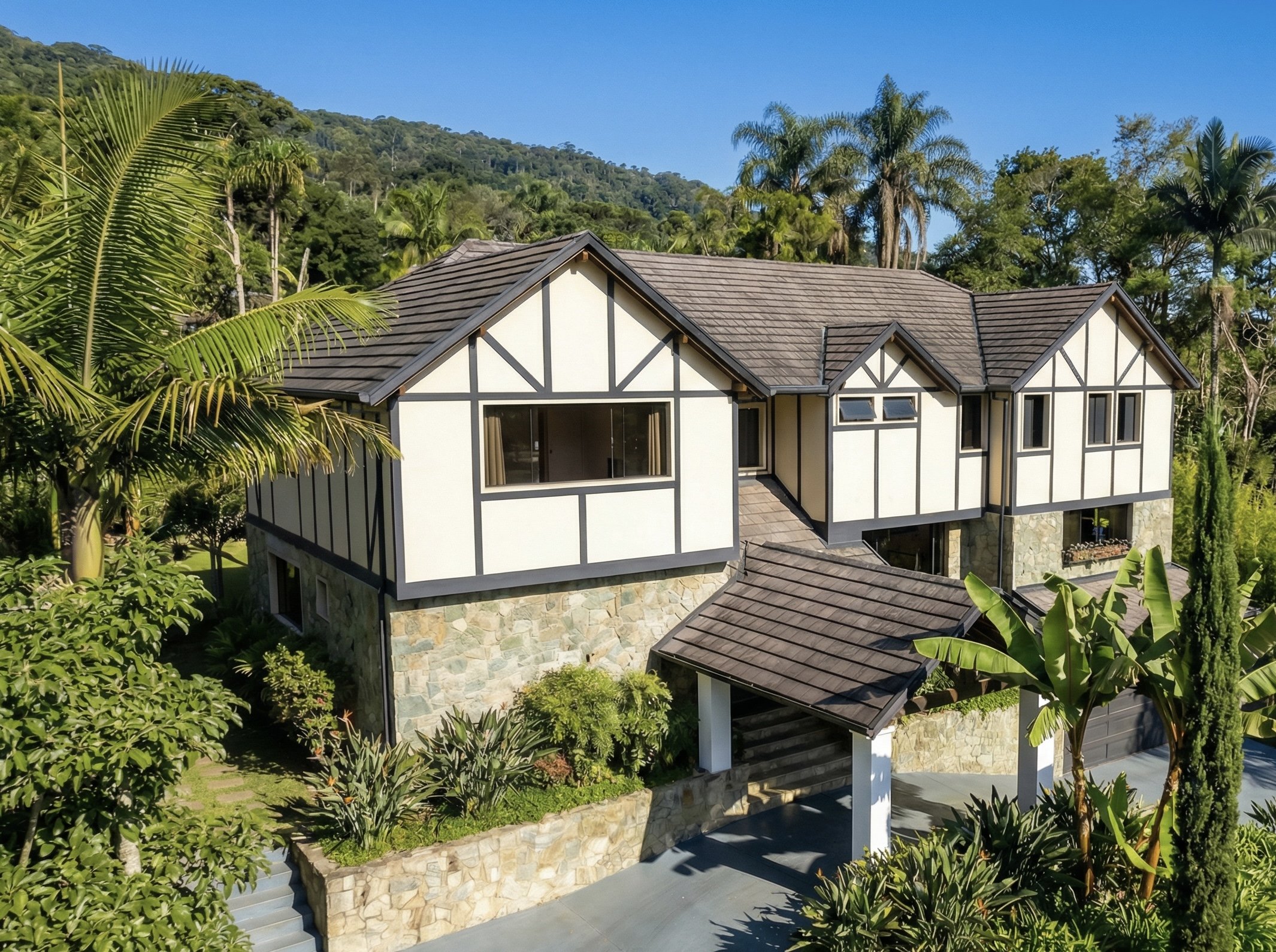 A modern two-story house with white walls and black timber framing, surrounded by lush green tropical plants and palm trees, with a mountain and clear blue sky in the background.