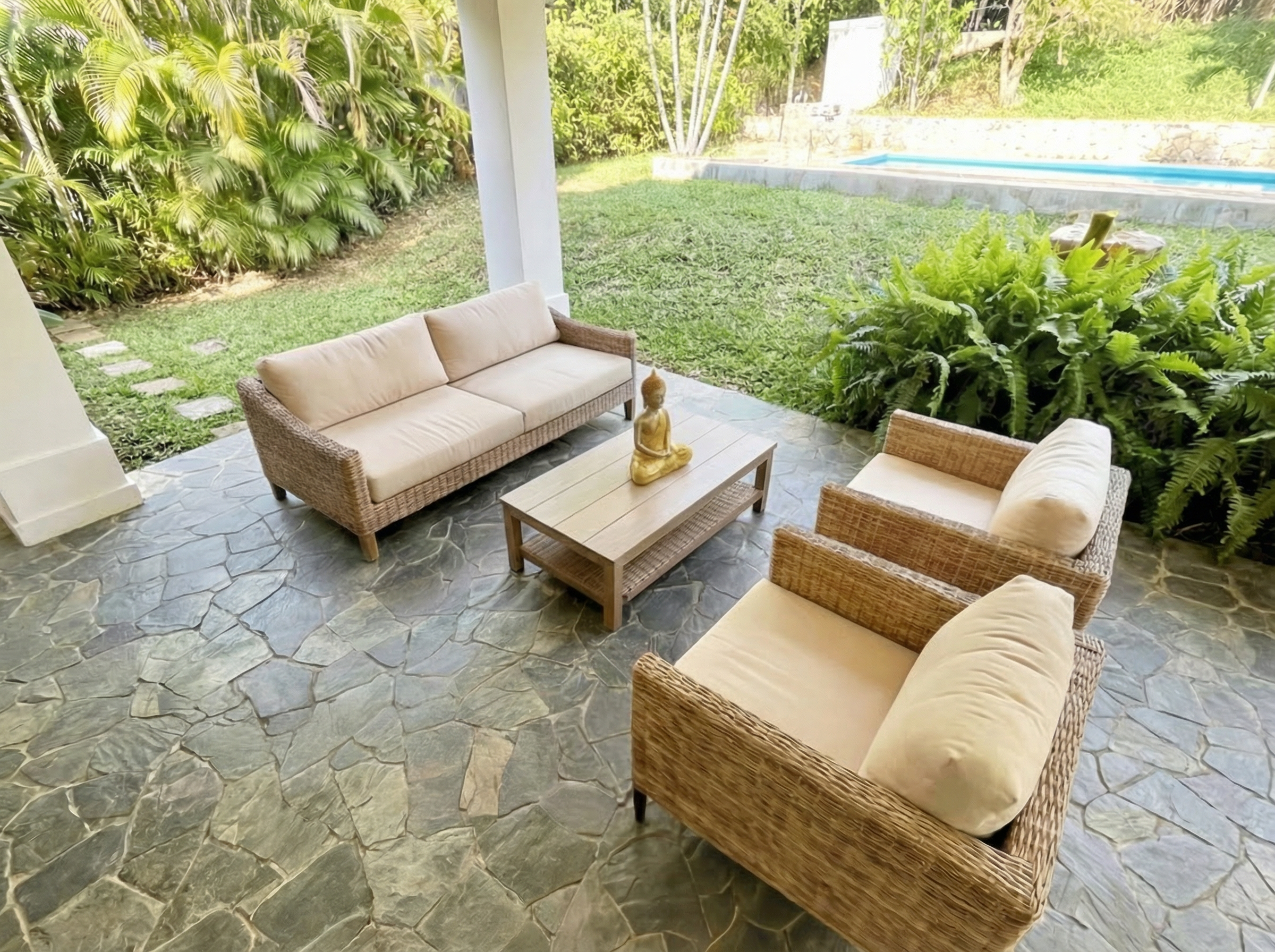 A patio area with wicker furniture including a beige sofa and two armchairs, a small wooden coffee table with a Buddha statue, surrounded by lush greenery, with a swimming pool in the background.