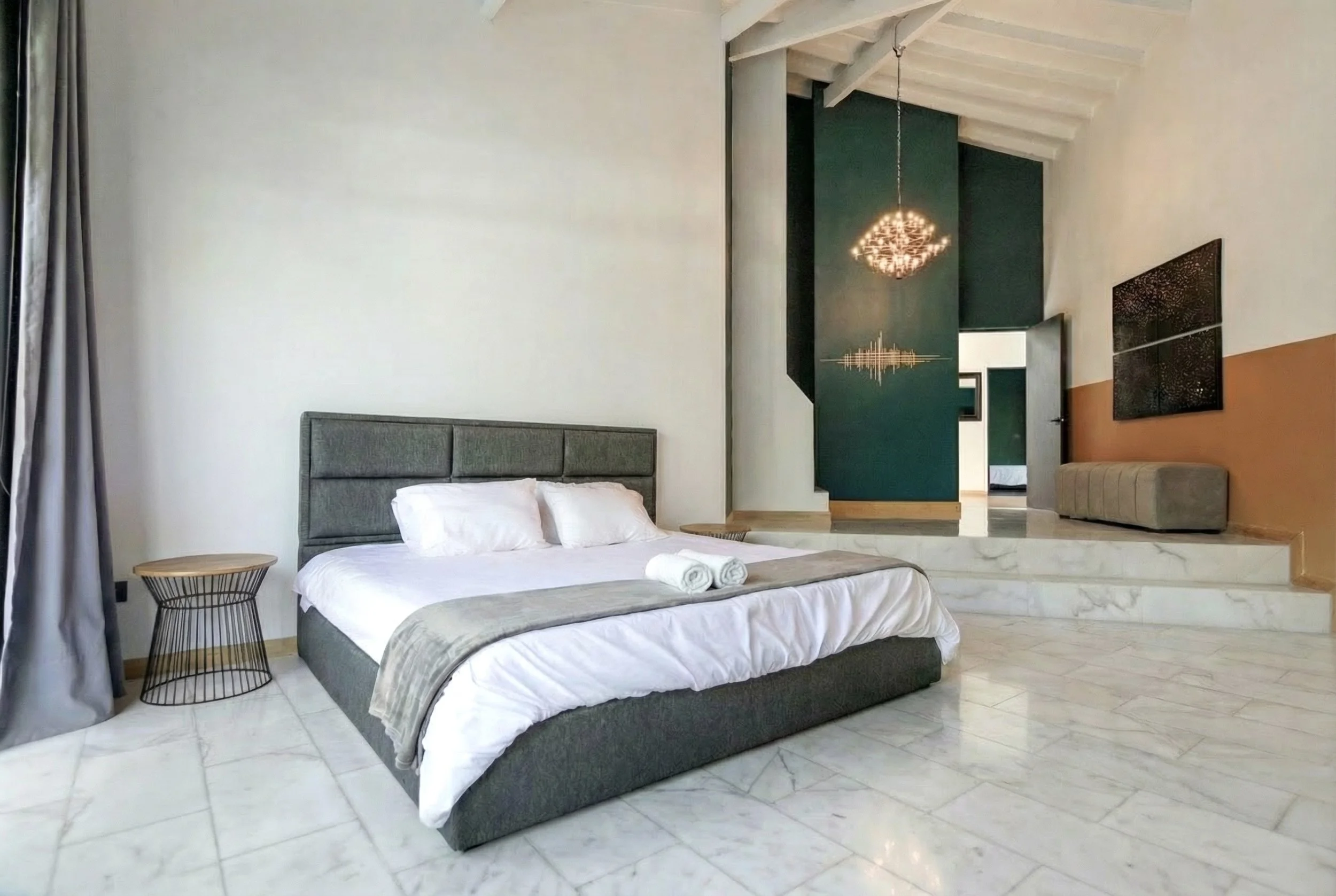 A modern bedroom with a large bed, gray upholstered headboard, white bedding, and rolled towels on the bed. There is a small round side table and gray curtains near a window on the left. The room features marble flooring and artistic decor on the wal
