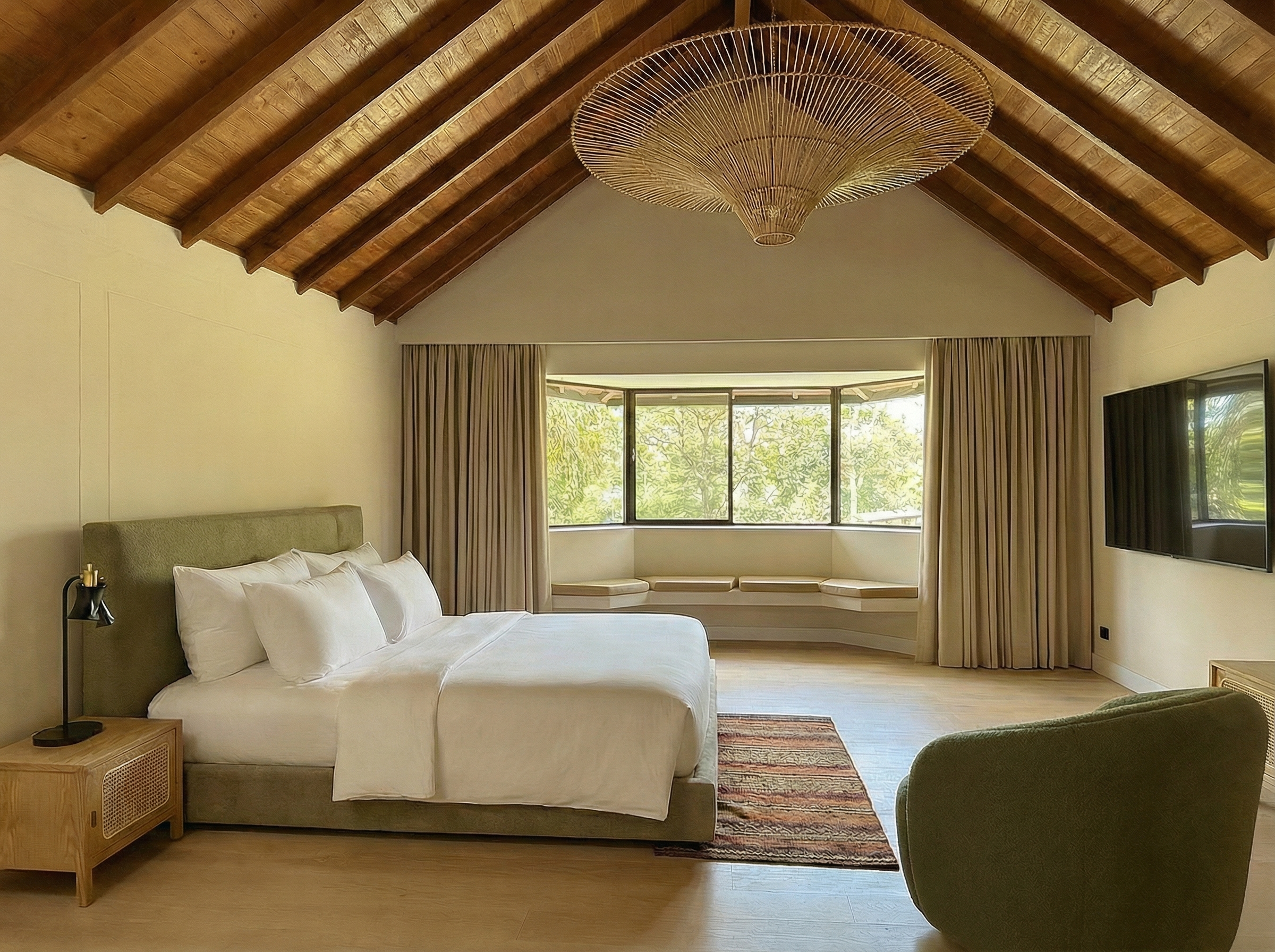 A bedroom with a wooden ceiling, a large bed with white bedding, a bedside table with a modern black lamp, a colorful rug, a curved window seat with beige cushions, and large windows with beige curtains showing green trees outside.