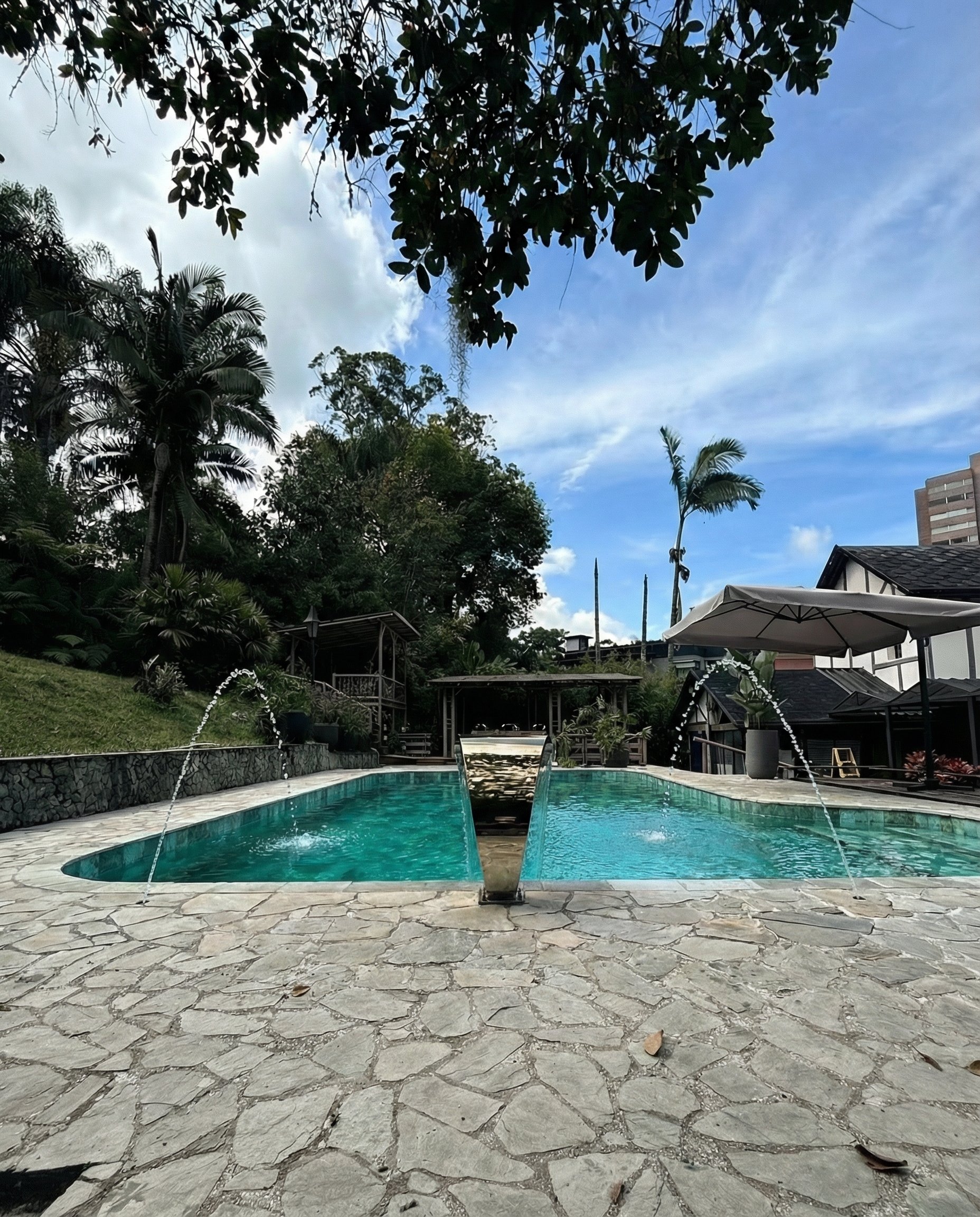 An outdoor swimming pool with clear water, surrounded by a stone patio. Tall trees and lush greenery are in the background. There is a large white umbrella on the right side, and a short fountain in the middle of the pool shooting water upwards. The 