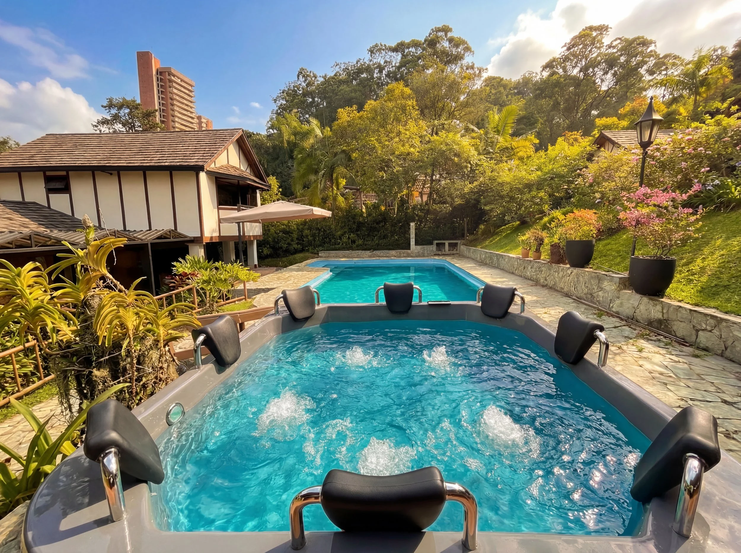 Hot tub with black headrests and metal handrails, surrounded by potted plants, next to a house with a tiled roof, overlooking a lush garden with trees and flowers, and a swimming pool, under a blue sky with scattered clouds.