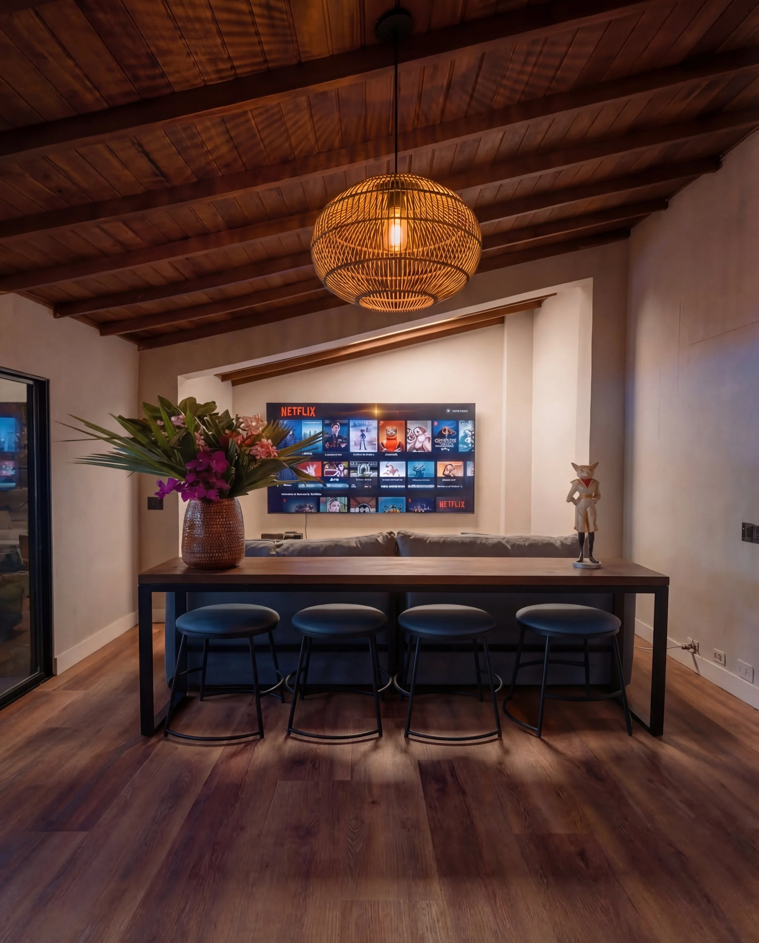 Living room with wooden ceiling, hanging pendant light, TV displaying Netflix interface, potted plant with pink flowers, and a wooden dining table with black chairs.