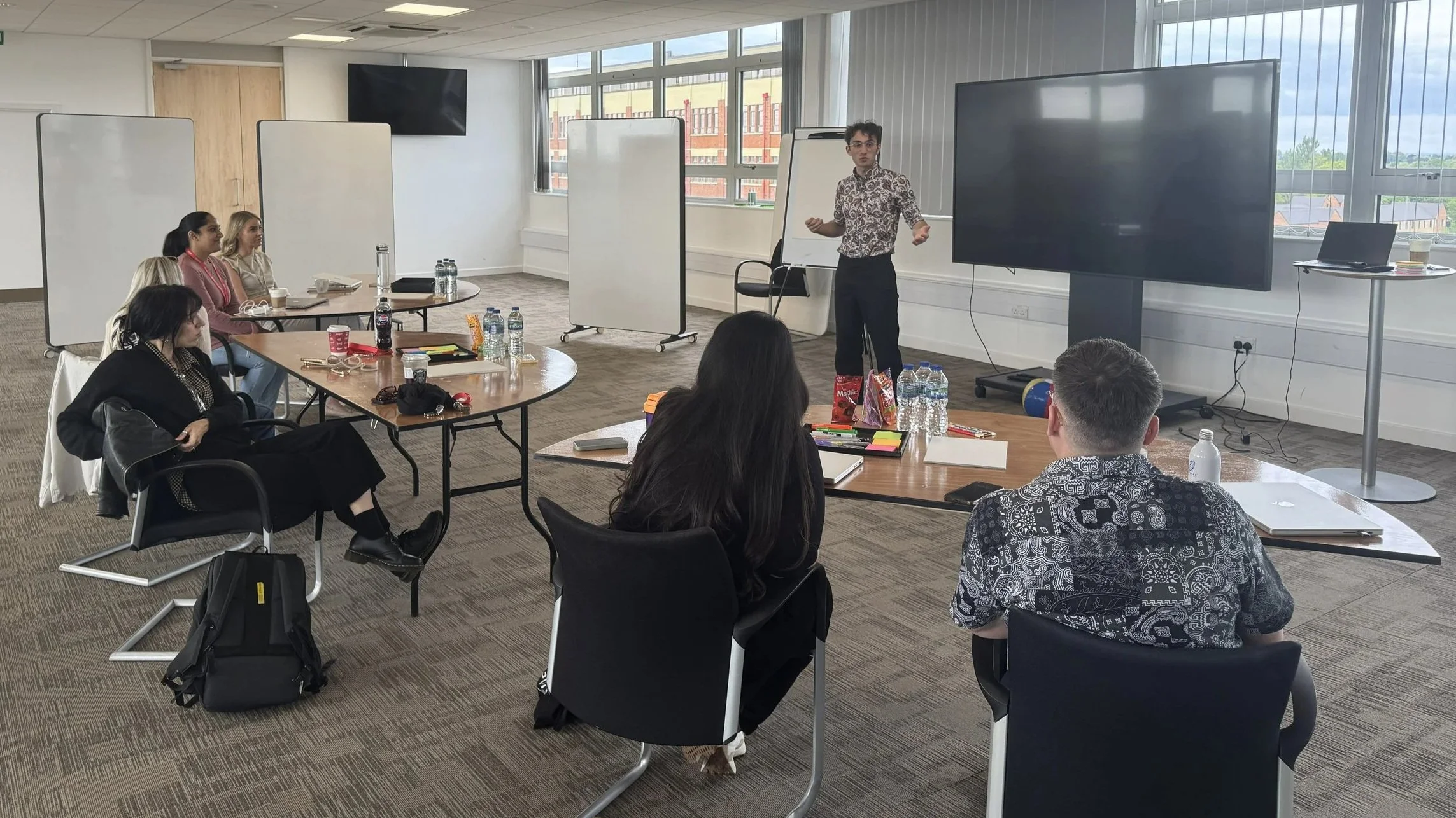 A man giving a presentation in a conference room with several attendees seated around tables, large windows, whiteboards, a large screen, and various water bottles and snacks on the tables.