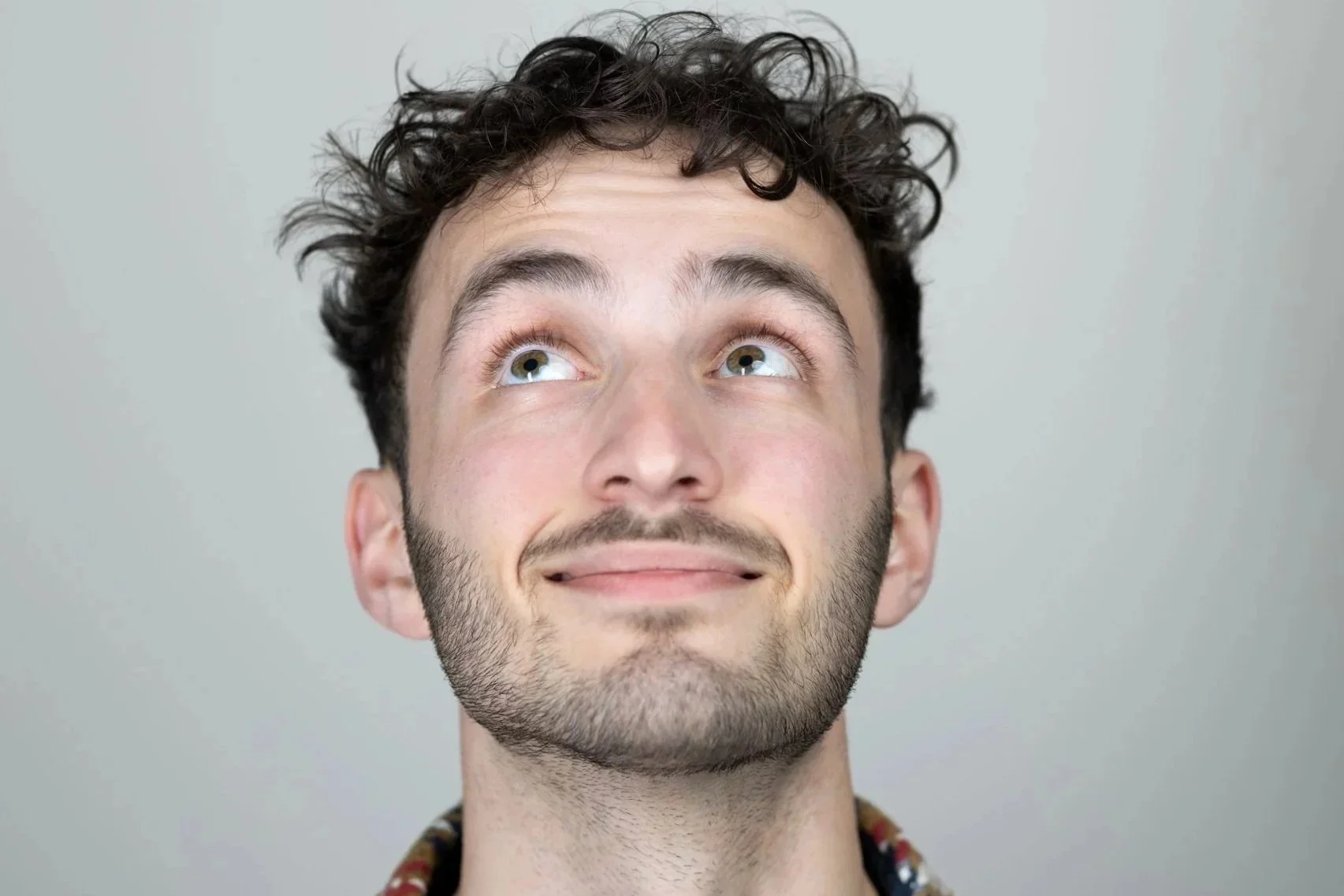 A young man with curly dark hair and a beard, smiling and looking up against a plain light gray background.