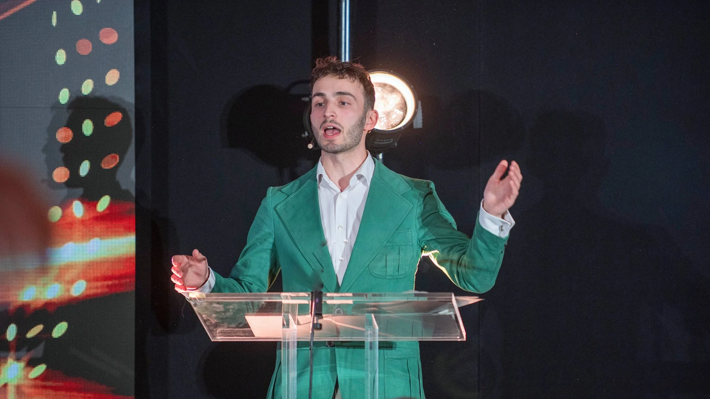 A man with short curly brown hair, wearing a white shirt and a teal blazer, speaking at a podium with a microphone. Behind him is a dark background with stage lighting and a large LED screen displaying colorful dot patterns.