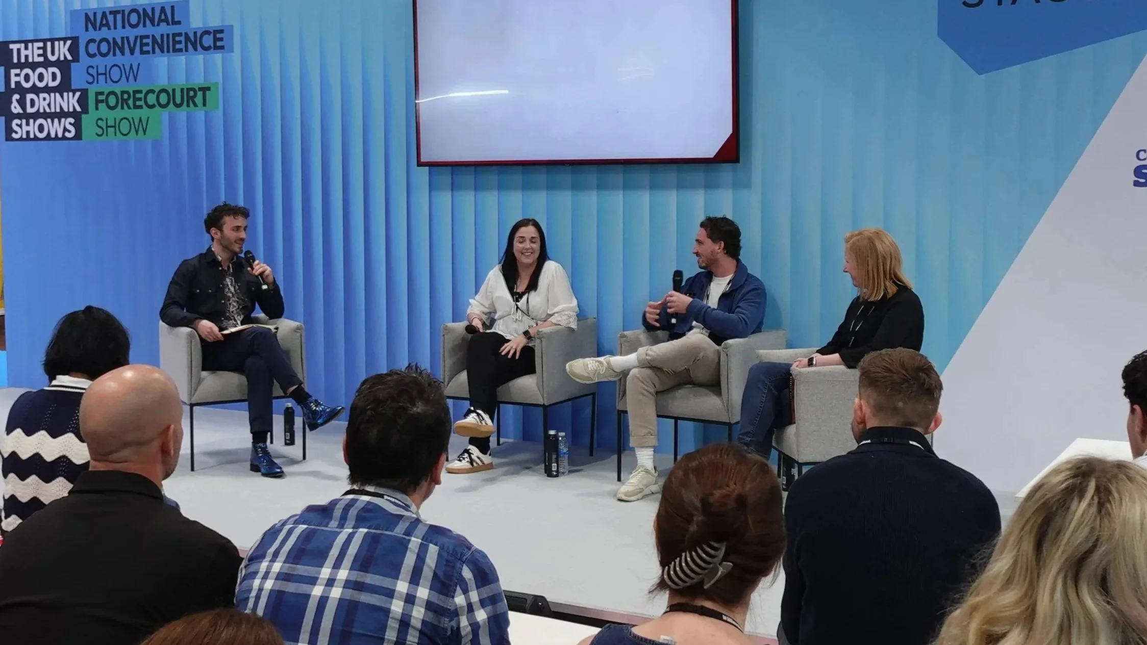 A panel discussion at a show titled 'The UK Food & Drink Show.' Four people are seated on stage in armchairs, engaged in conversation. An audience is sitting in front of them, watching the panel.
