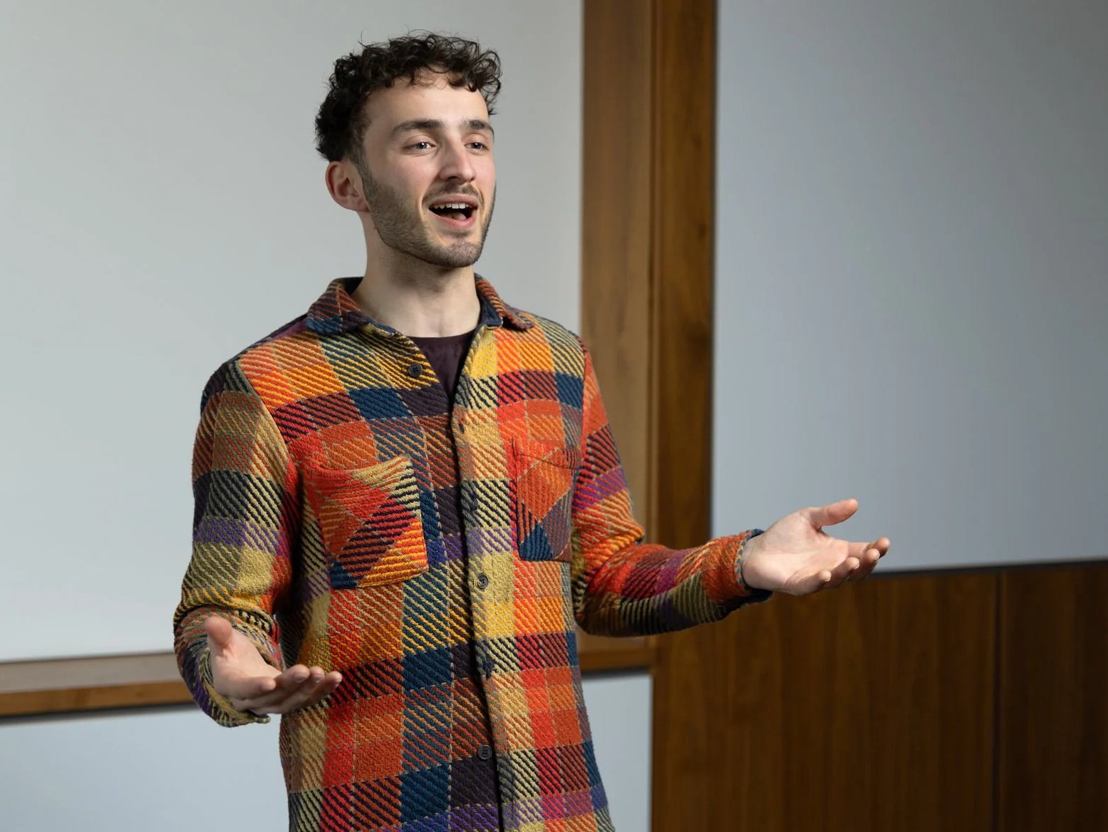 A man with curly dark hair and a beard, wearing a colorful plaid shirt, appears to be speaking and gesturing with open hands in a room with wooden panels and a whiteboard.
