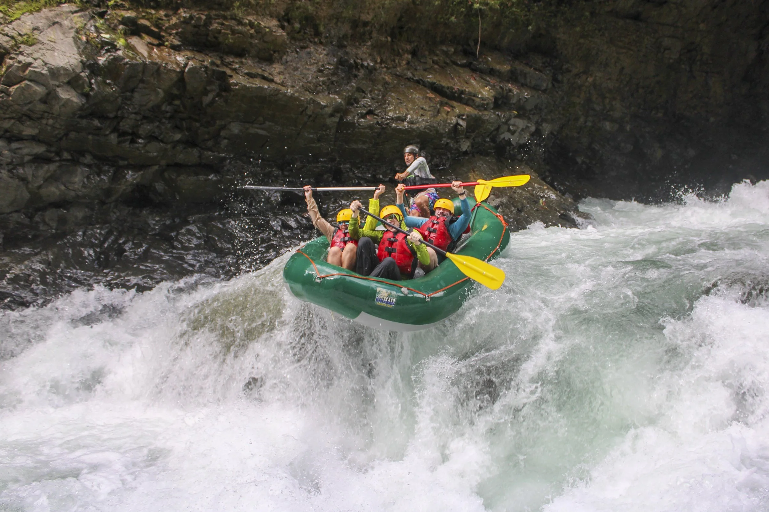 Four people in a green inflatable raft, wearing life jackets and helmets, navigate rapids in a river, guided by a person standing at the back with a paddle.