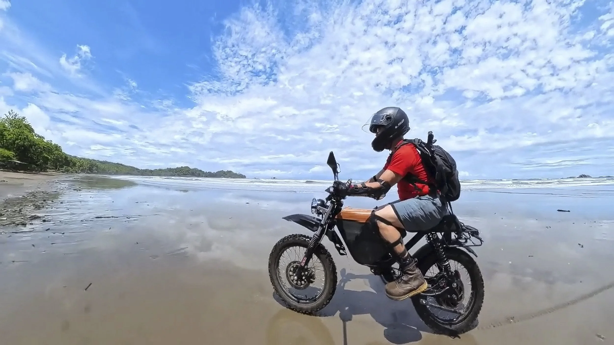 A person riding a black electric dirt bike on a sandy beach with water and sea waves in the background, under a partly cloudy sky.