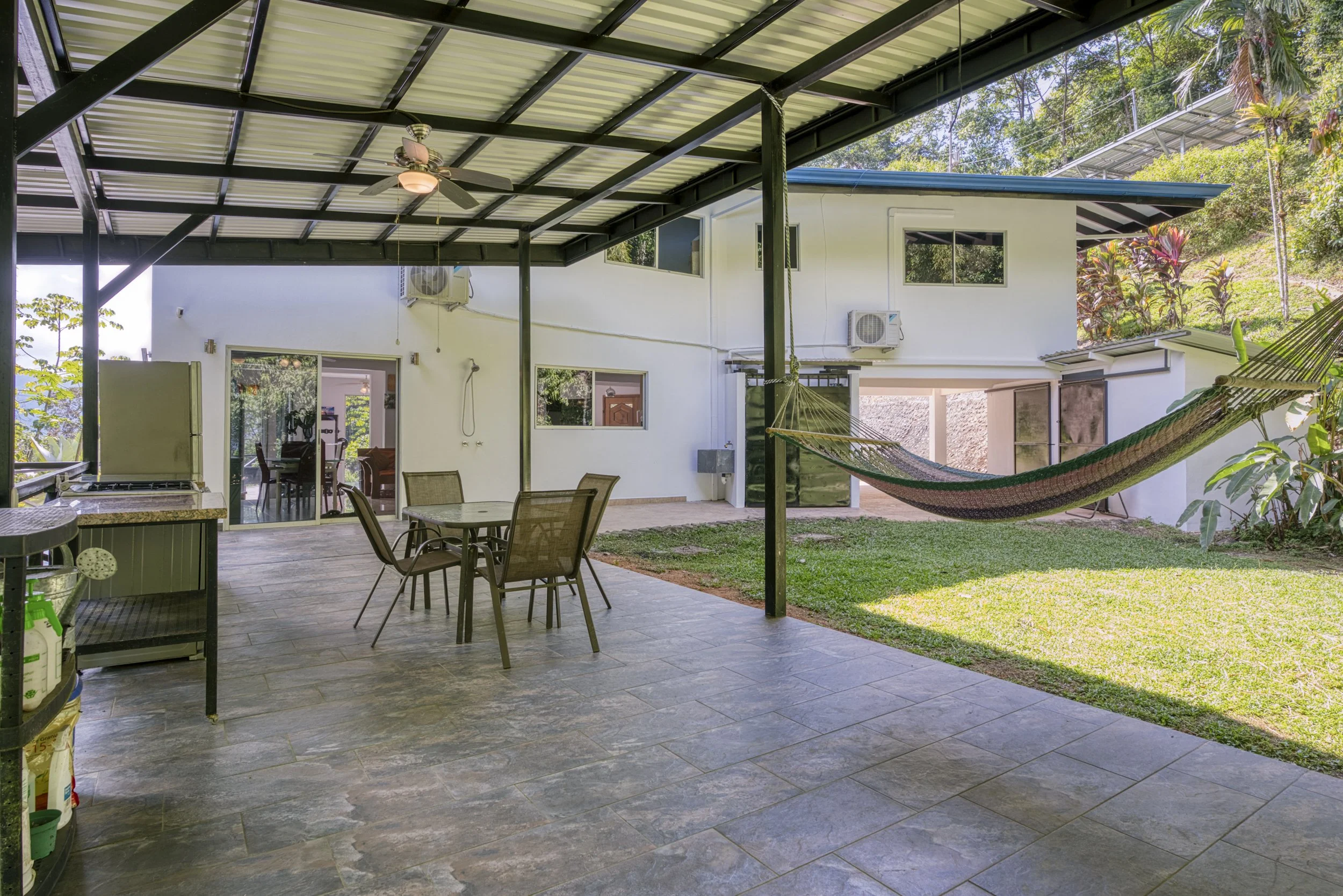Covered patio area with a ceiling fan, outdoor table with four chairs, hammock tied to support poles, and a grassy yard with tropical plants in the background.