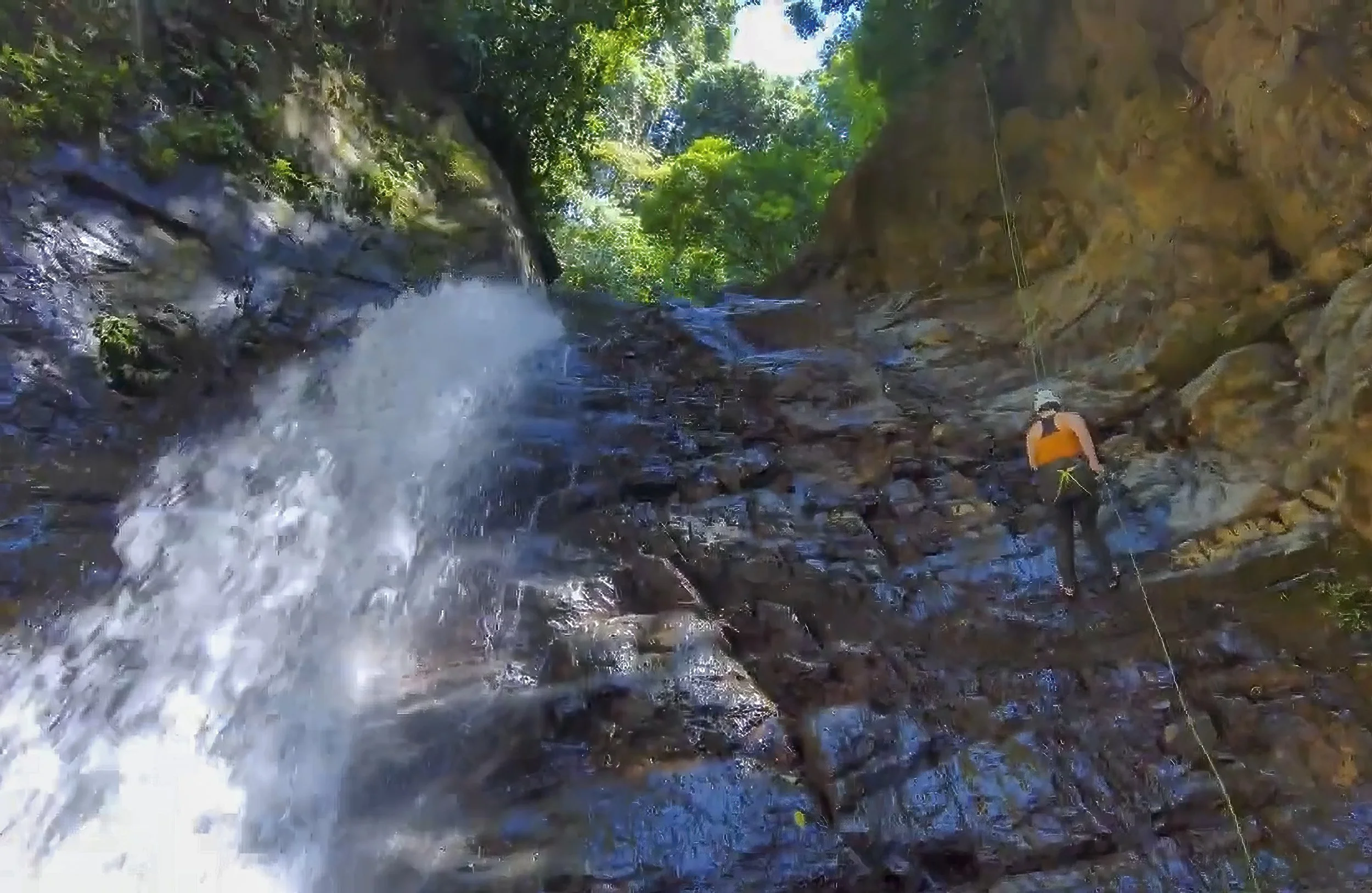 A person rappelling down a rocky waterfall in a forested area during daytime.
