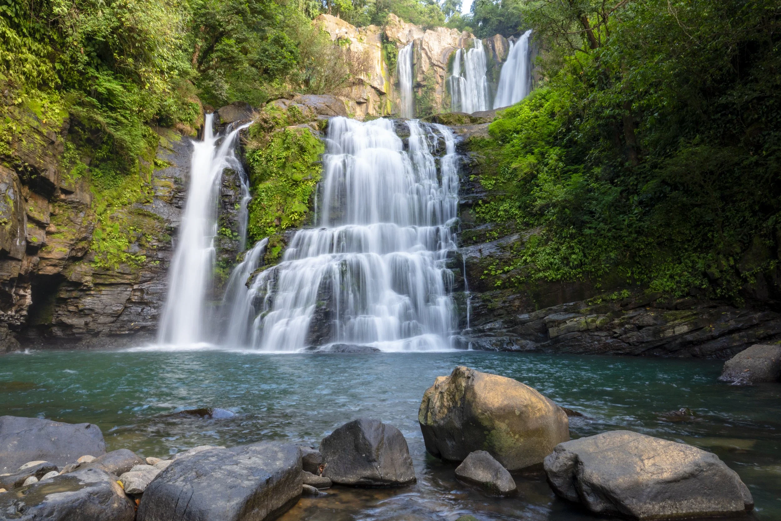 A multi-tiered waterfall flowing into a pool surrounded by rocks and lush green trees.