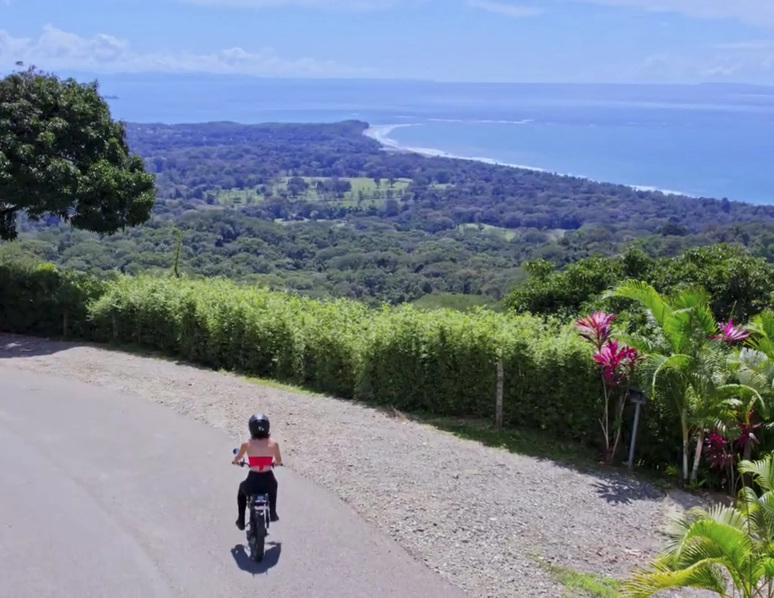 A young girl riding a bicycle on a paved road overlooking a lush, green landscape with hills, trees, and the ocean in the distance. She is wearing a helmet and a red top.