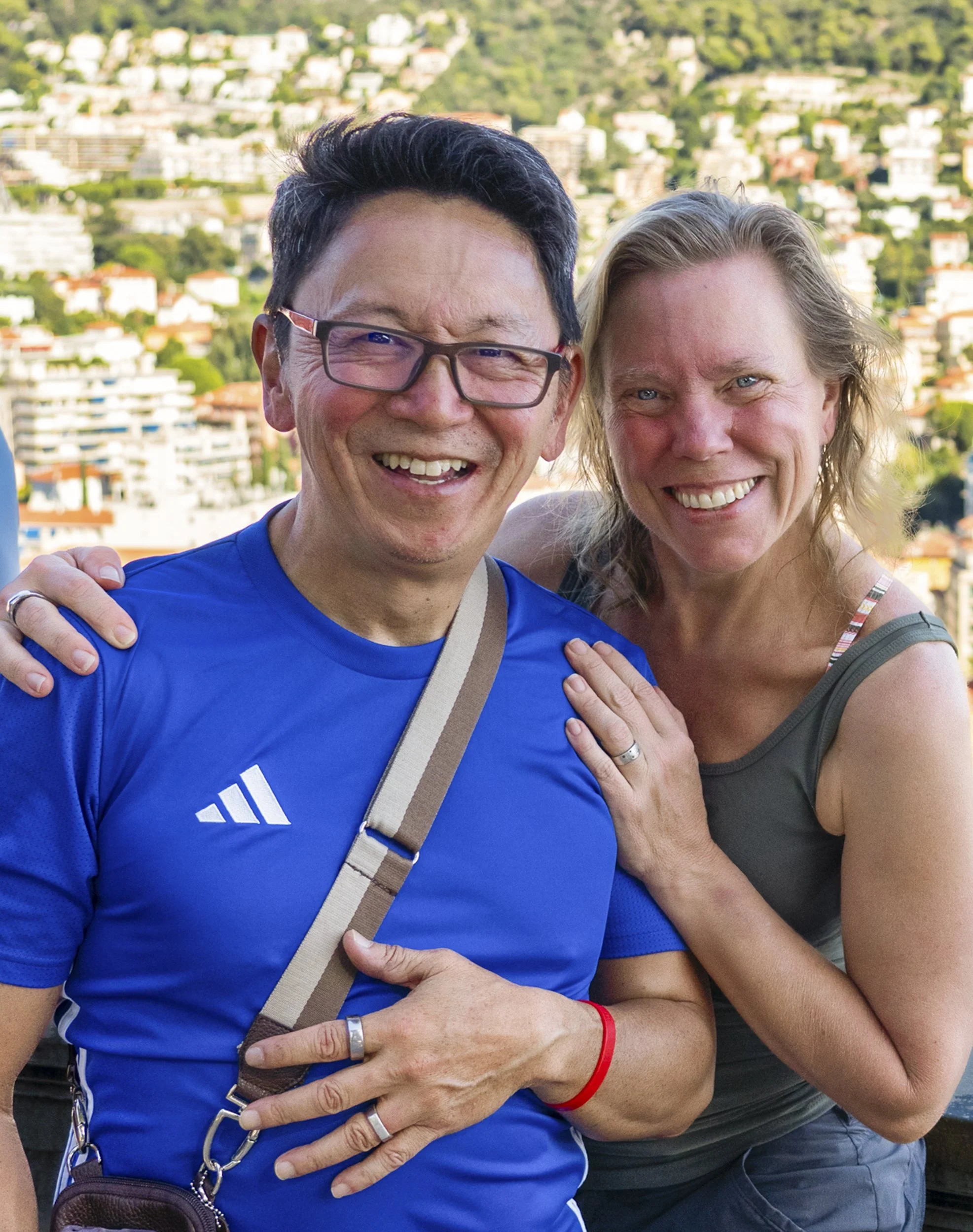 Two smiling people, a man wearing glasses and a blue shirt and a woman with wavy blonde hair and a tank top, pose together outdoors with a cityscape and green hills in the background.