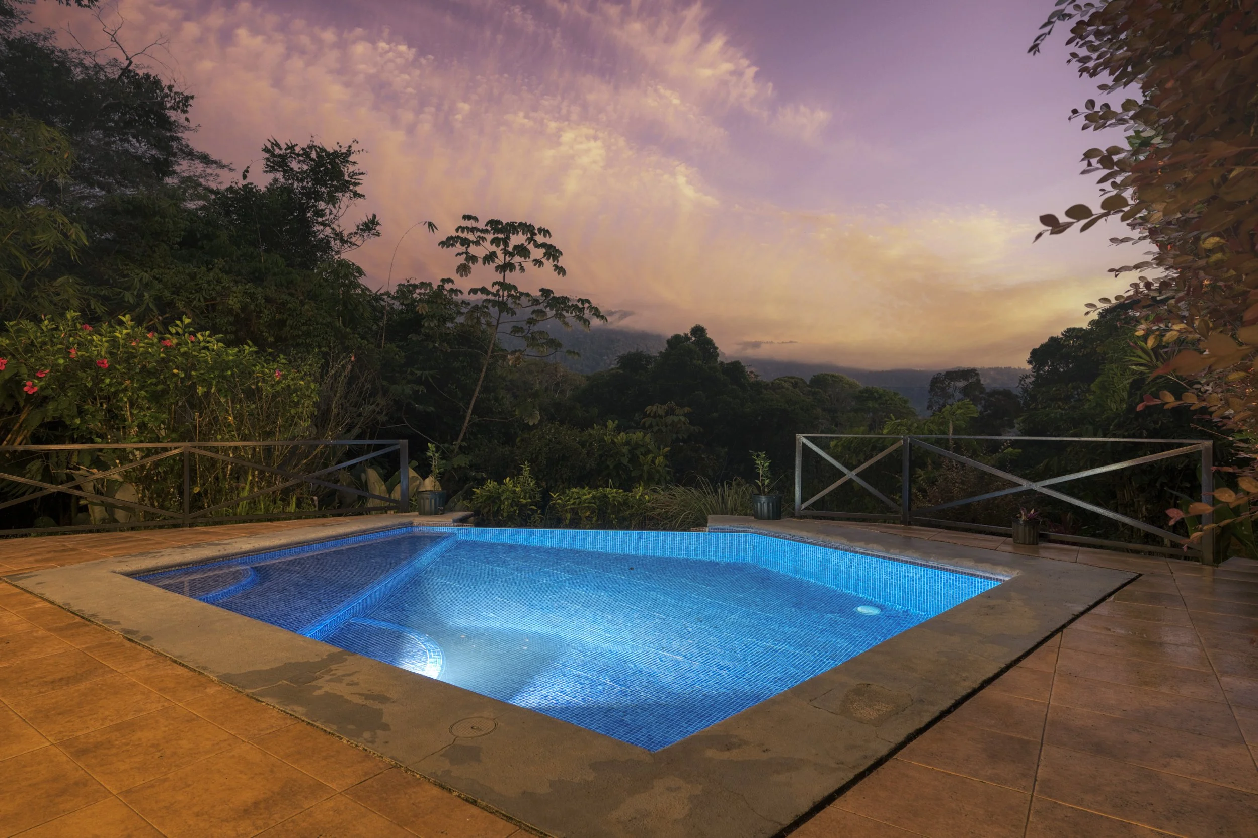 A swimming pool with blue lighting, surrounded by a tiled deck, overlooking a mountain landscape at sunset with colorful clouds in the sky.